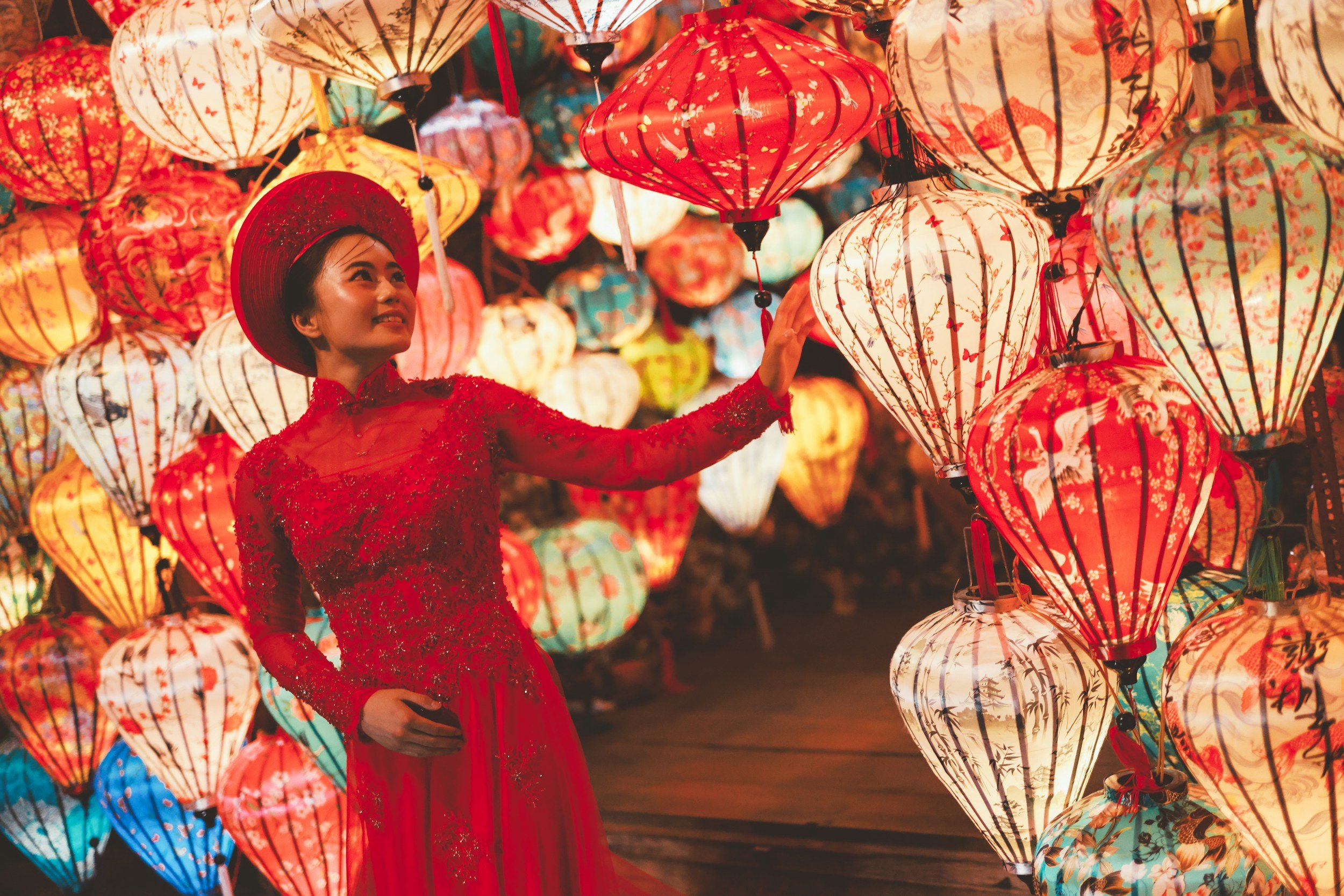 A woman in a red lace dress and matching hat touching colorful paper lanterns at night.