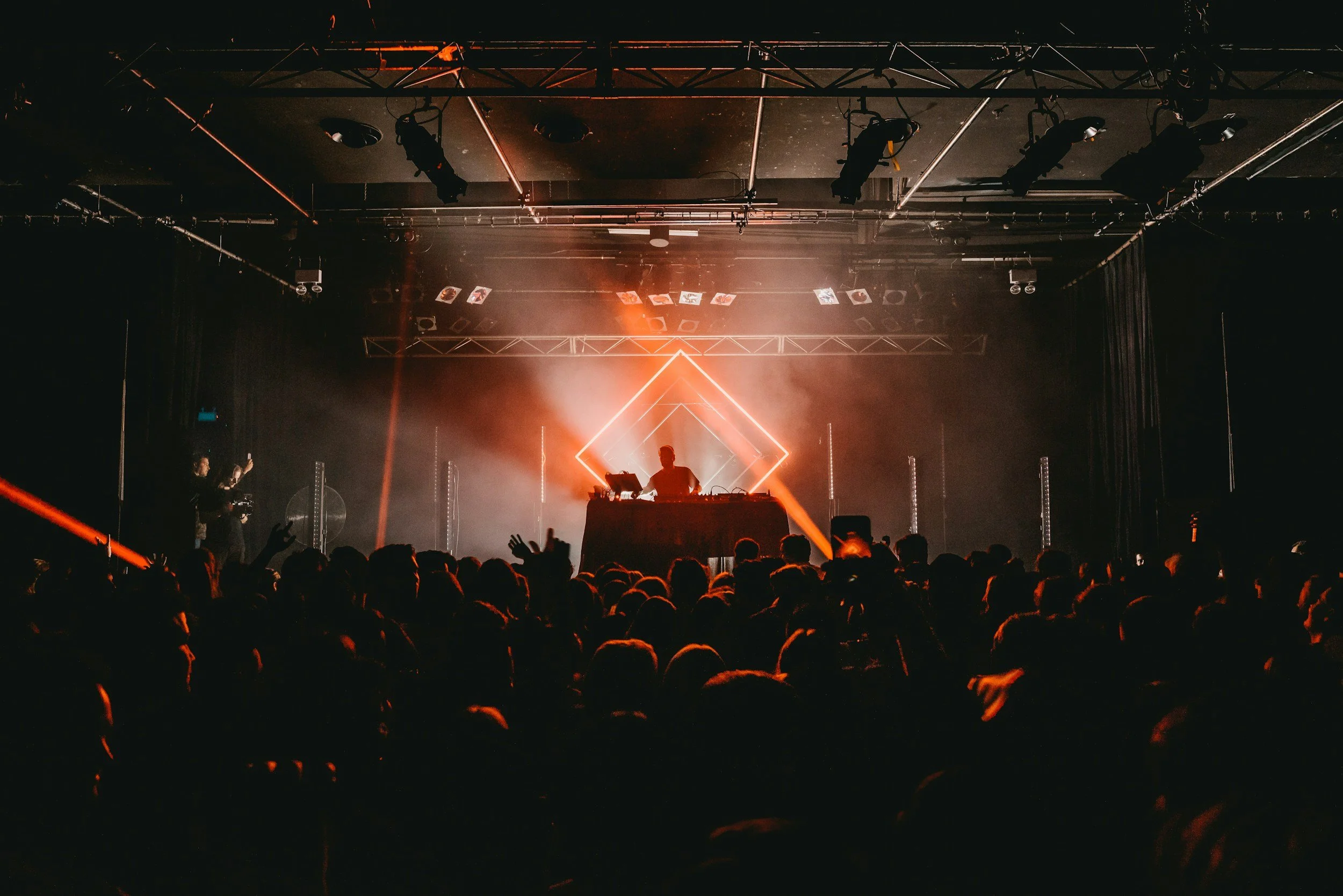 A DJ performing on stage at a concert with a crowd in front, illuminated by orange lighting, with geometric light patterns and stage equipment overhead.