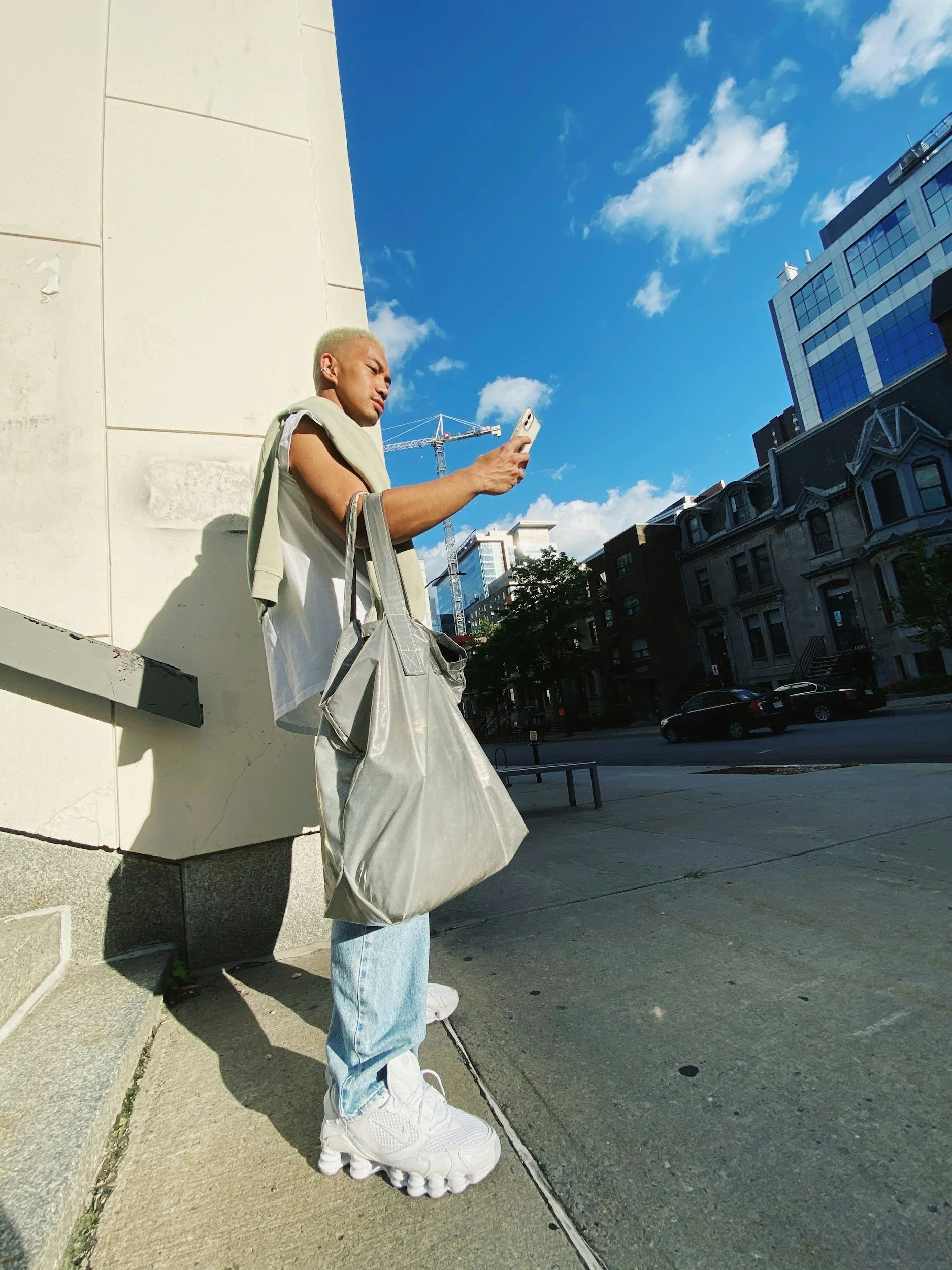 A young man with bleached blonde hair is standing on a city sidewalk, looking at his smartphone. He's dressed in a white hoodie, light blue jeans, and chunky white sneakers. He's carrying multiple bags, including a large gray tote. The background includes modern buildings, a street with parked cars, and a bright blue sky with a few clouds.