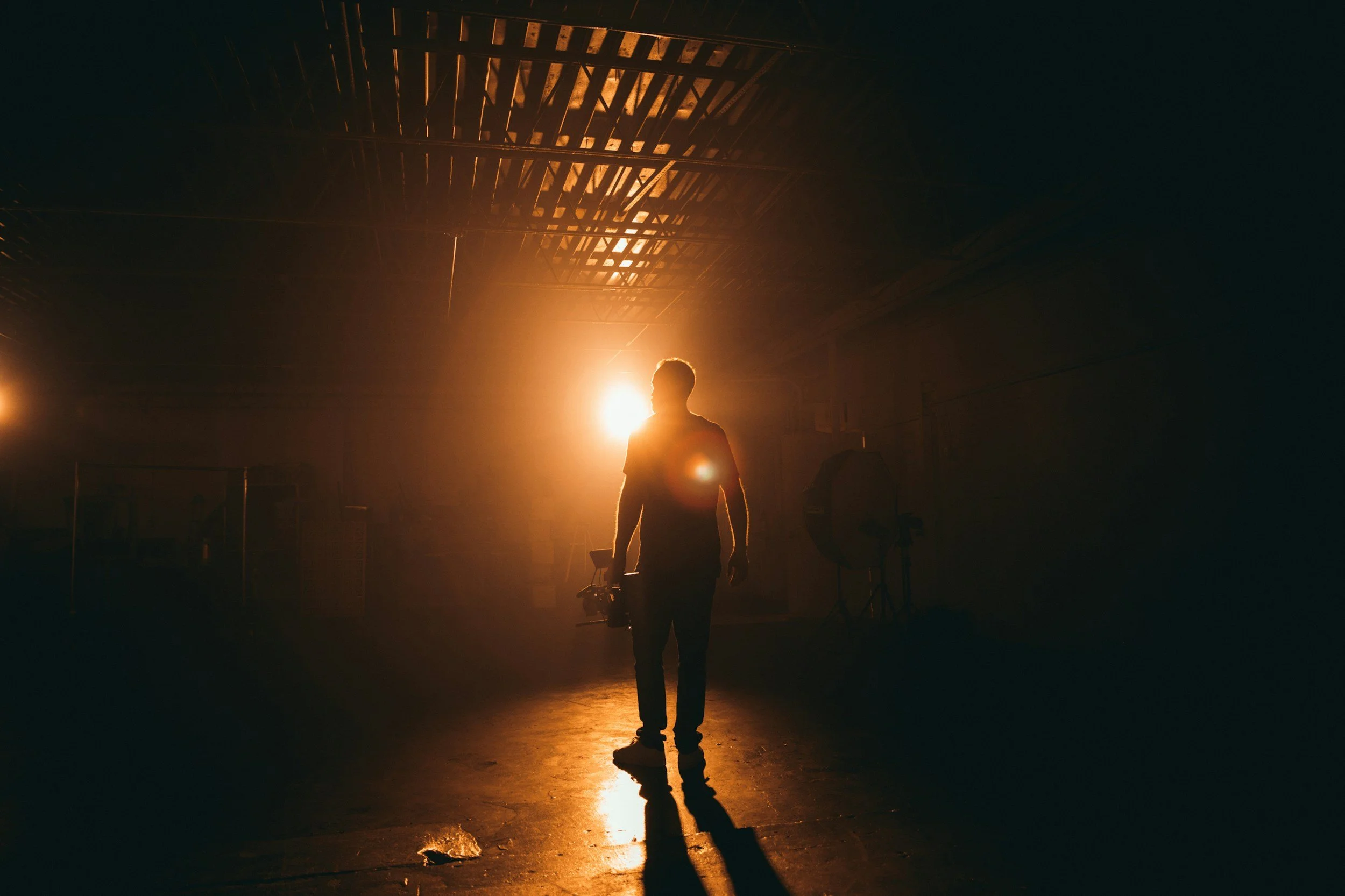 A person walking in an indoor space with dramatic lighting, backlit by a bright light source creating a silhouette effect, with a visible reflection on the floor.
