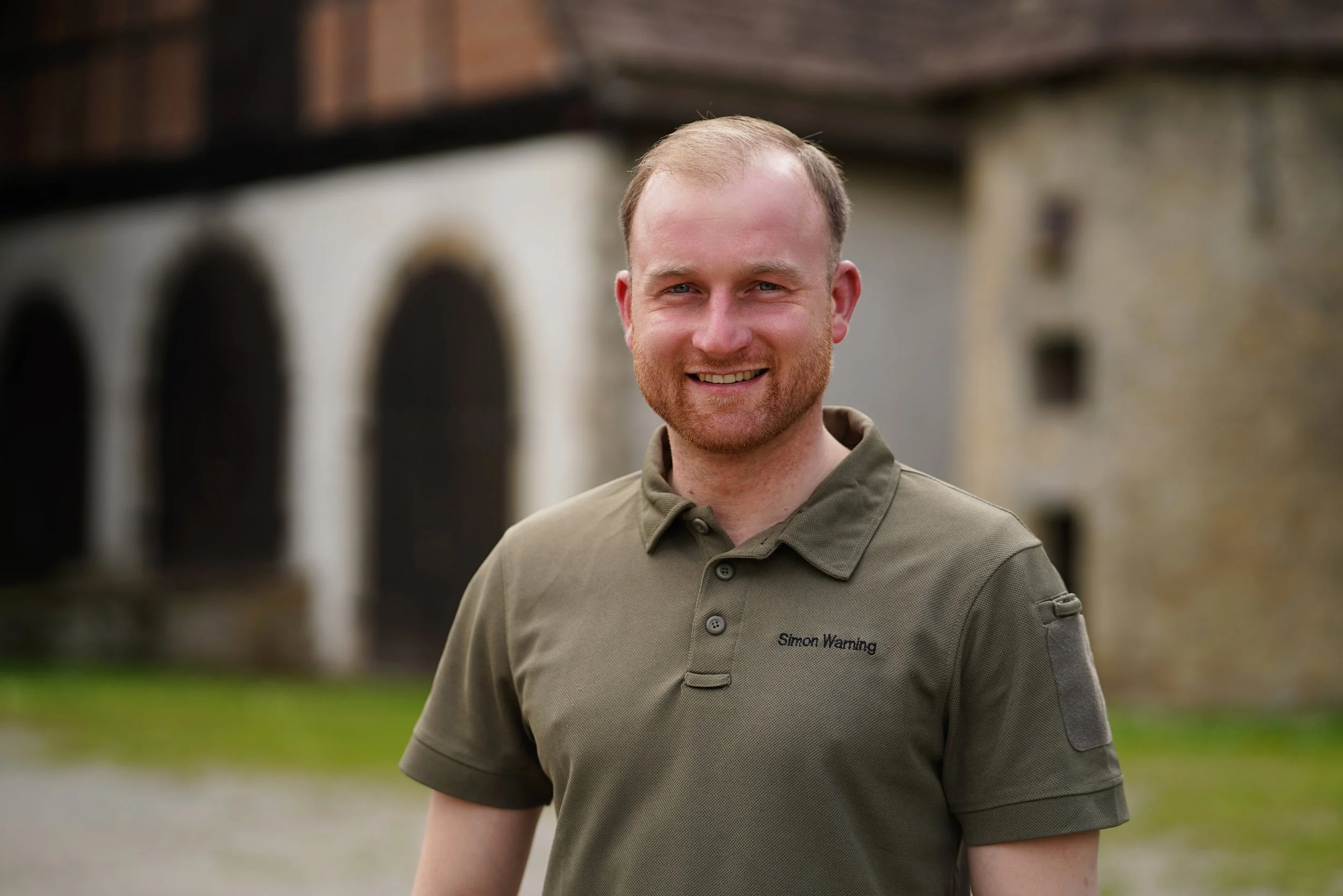 Ein Mann trägt ein khakifarbenes Polo-Shirt mit Aufdruck 'Simon Warming' auf der Brust, steht draußen vor einer alten Steinmauer und lächelt. HOB Wald und Naturraummanagement.