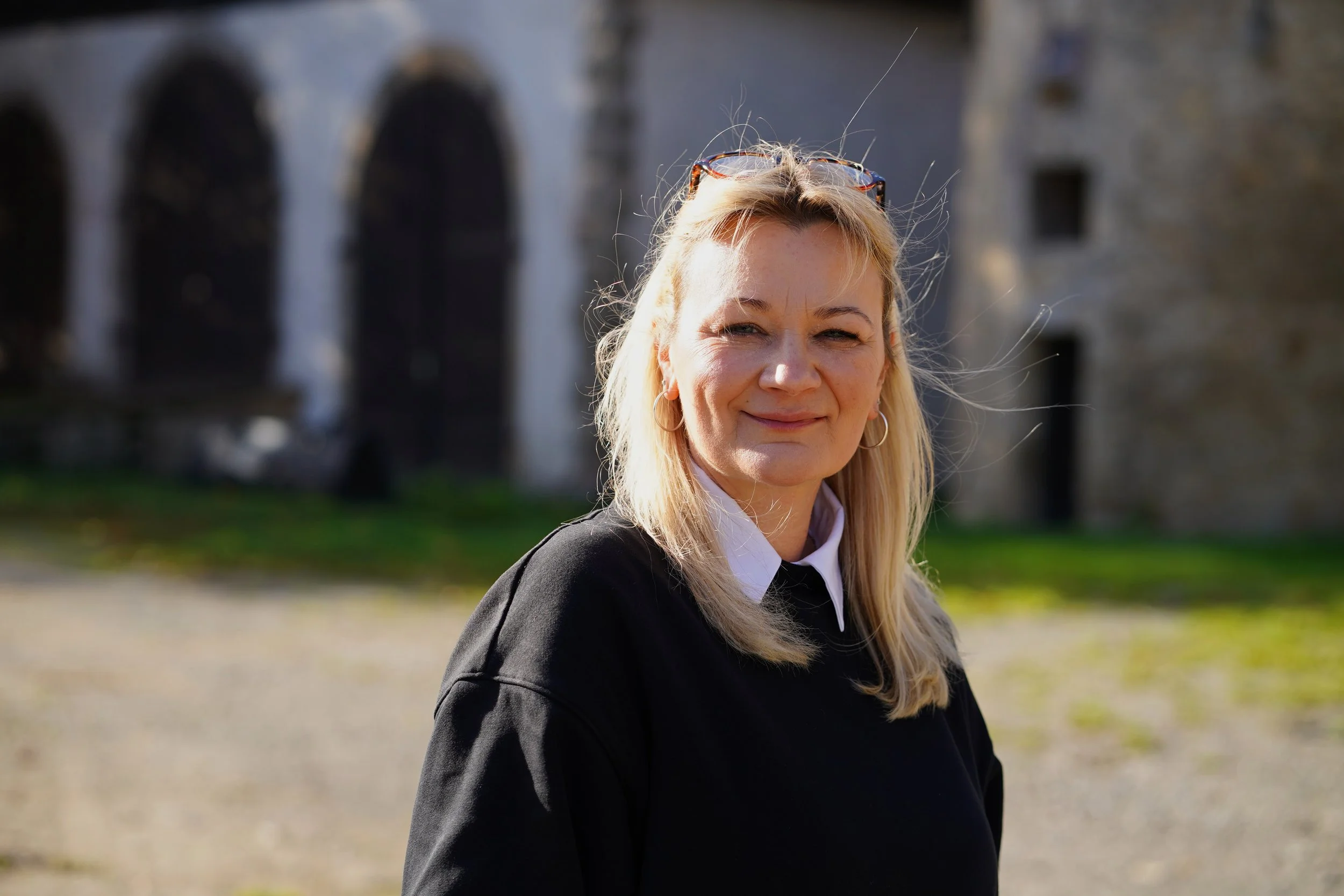 Frau mit langen blonden Haaren, Brille auf Kopf, lachend, draußen vor einer alten Steinmauer bei Sonnenlicht. Klaudia Piechaczek, AveNATURA, Naturbestattungen Gut Holzhausen, OWL, Kreis Höxter.