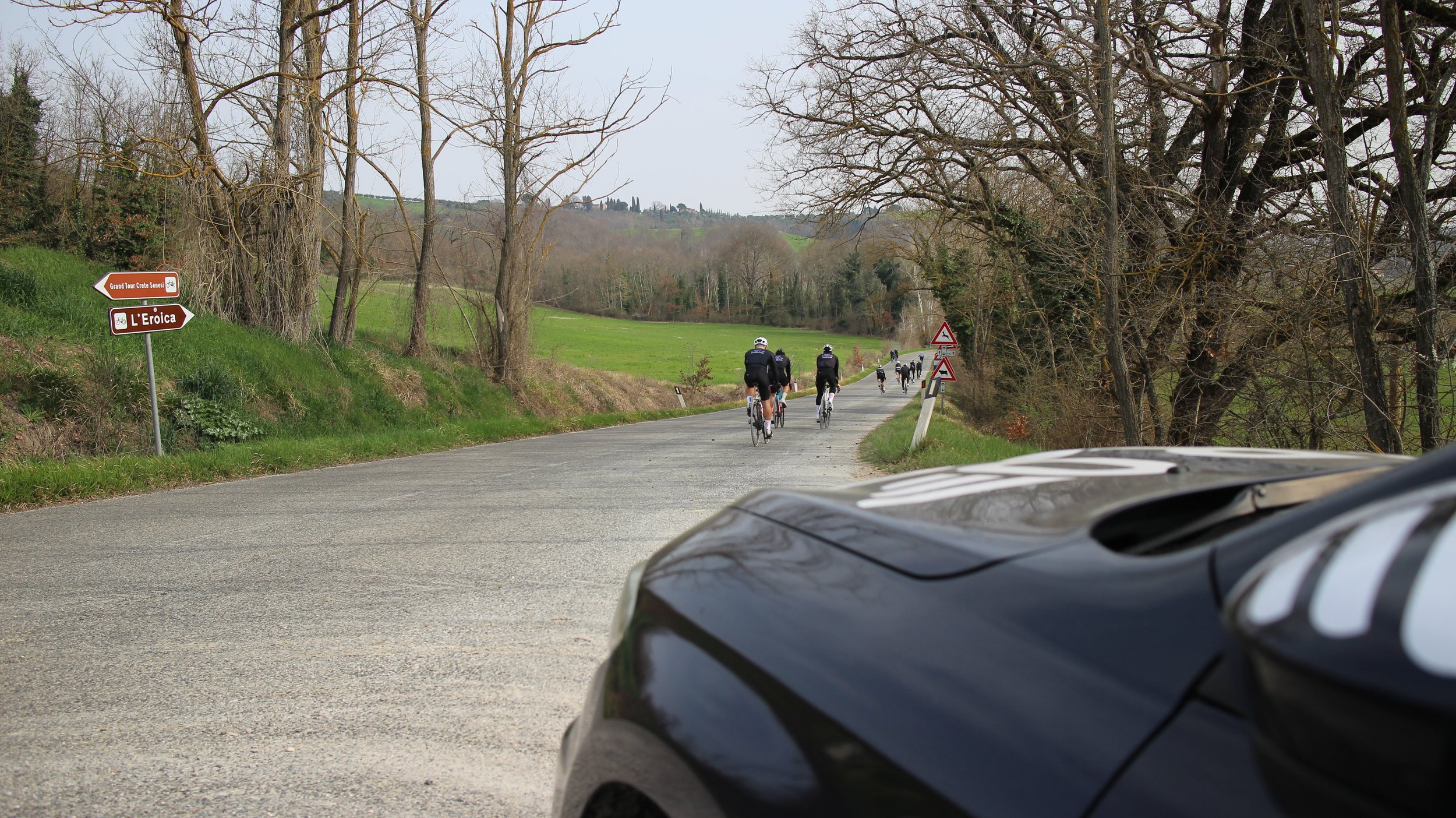Grupo de ciclistas pedalando por uma estrada rural cercada de árvores, com sinais de trânsito ao lado e colinas ao fundo, em uma paisagem de clima nublado.