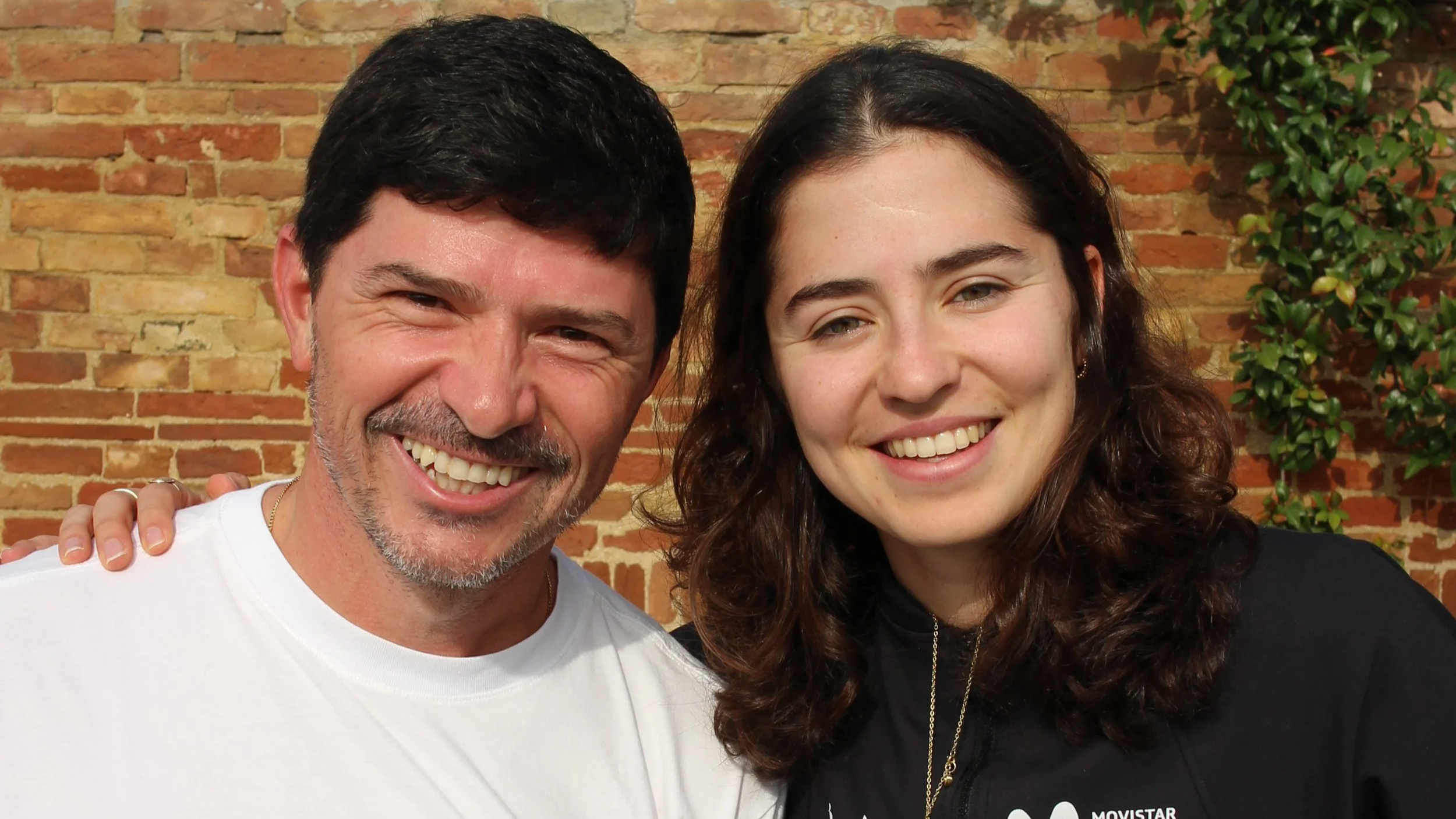 Homem e mulher sorrindo, o homem segura capacete de ciclismo e a mulher usa jaqueta com logo da Movistar, ao fundo parede de tijolos, árvores e céu azul.