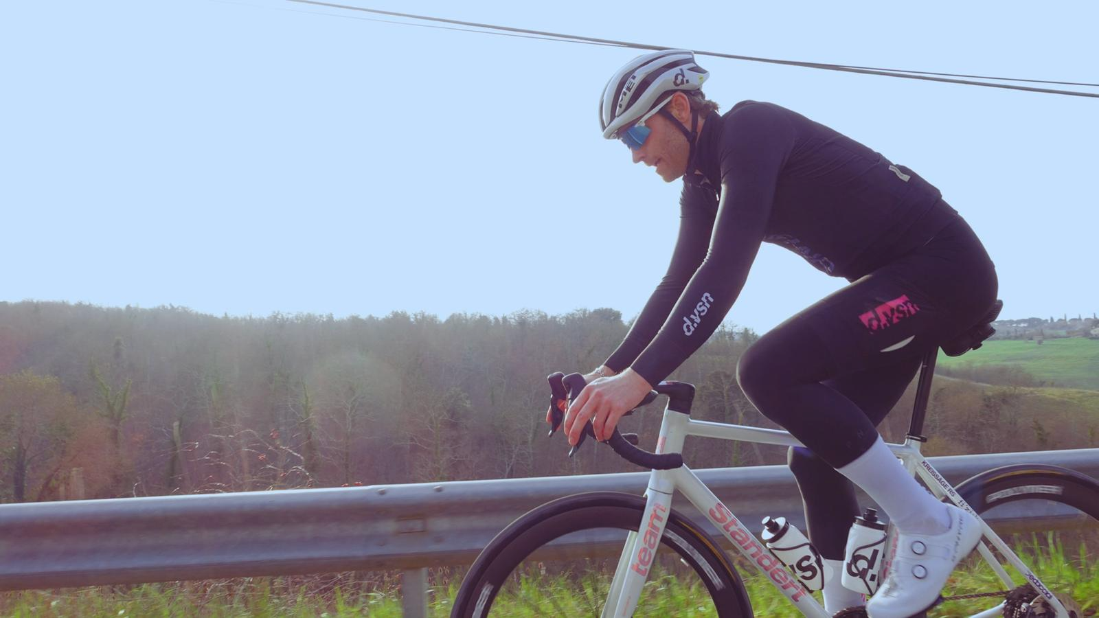 Ciclista de uniforme preto e branco pedalando na estrada com céu claro e campo ao fundo.
