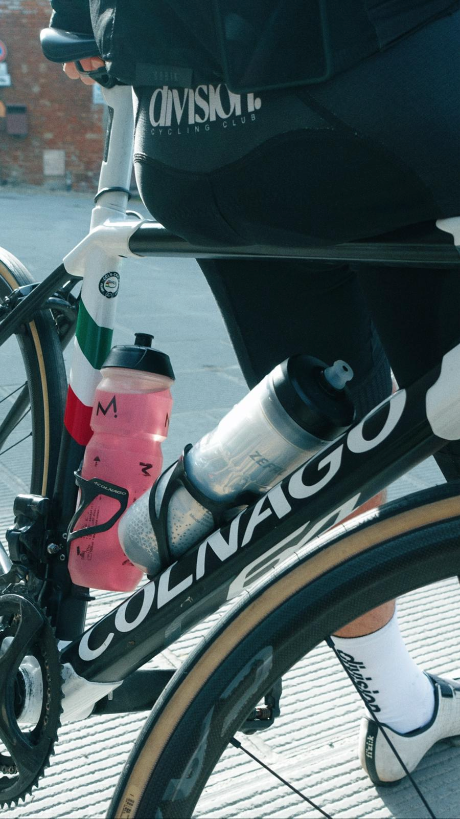 Parte de uma bicicleta de ciclismo com dois porta garrafas, uma rosa e uma prateada, com uma roda de estrada de cor preta e detalhes em marrom. Uma pessoa, vestindo roupas de ciclismo, está sobre a bicicleta, com a perna vestida com uma meia branca q