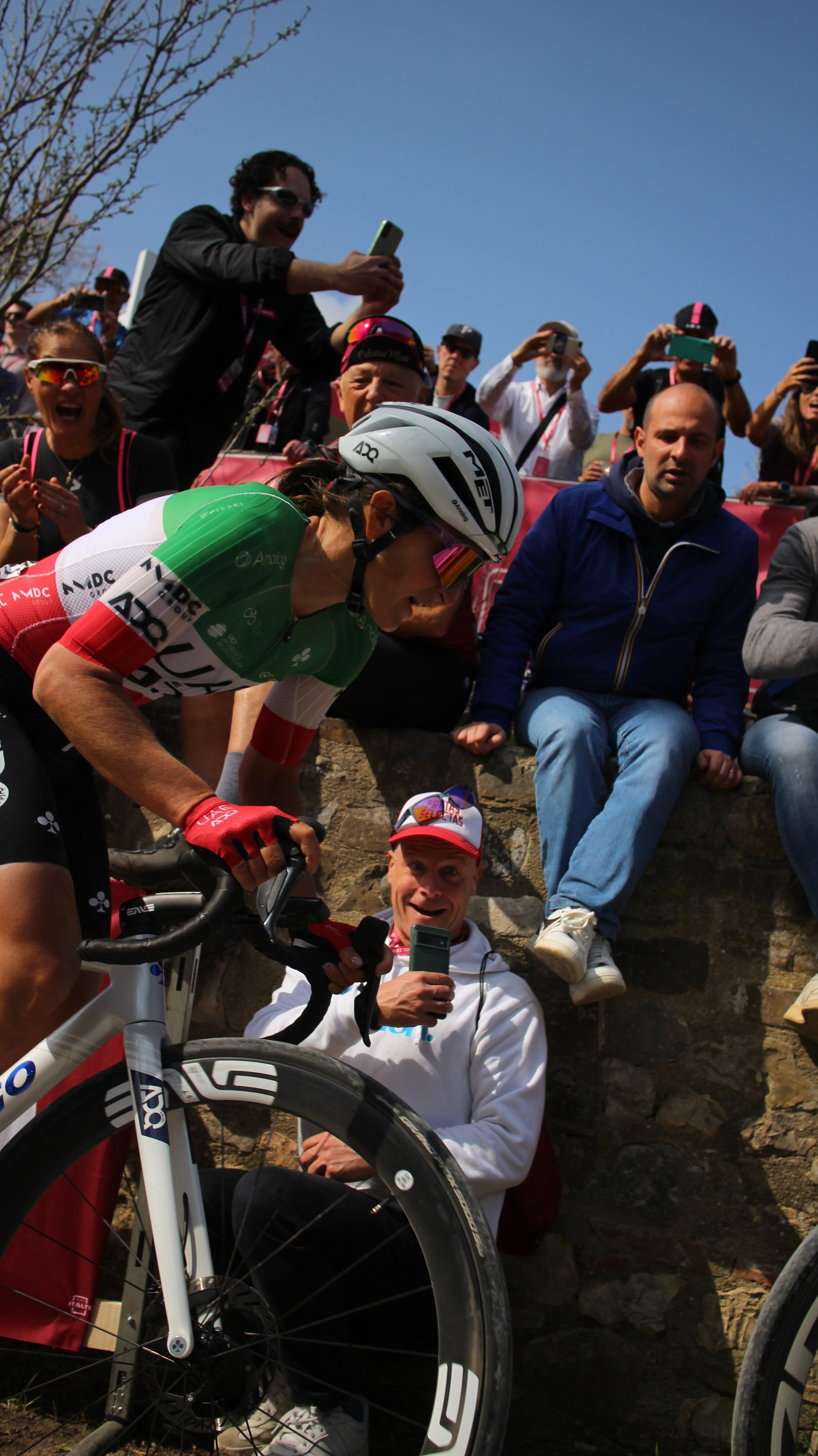 Ciclismo durante uma competição com um ciclista usando capacete e uniforme de time, rodeado por espectadores tirando fotos e vídeos, com o céu ao fundo.