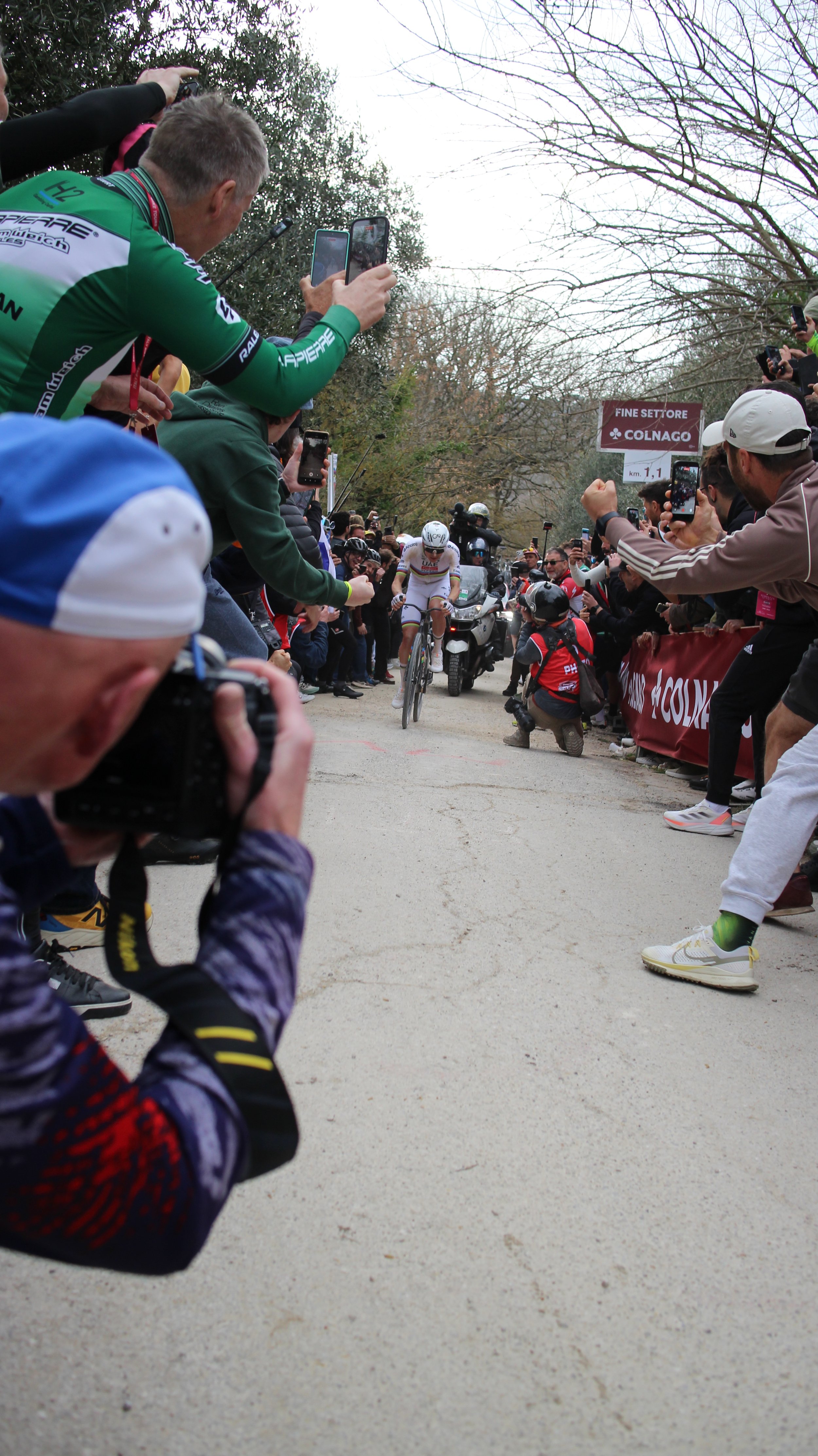 Ciclista em uma corrida sendo fotografado por várias pessoas com celulares ao redor, árvores sem folhas ao fundo e uma placa com o nome 'Colnago'.