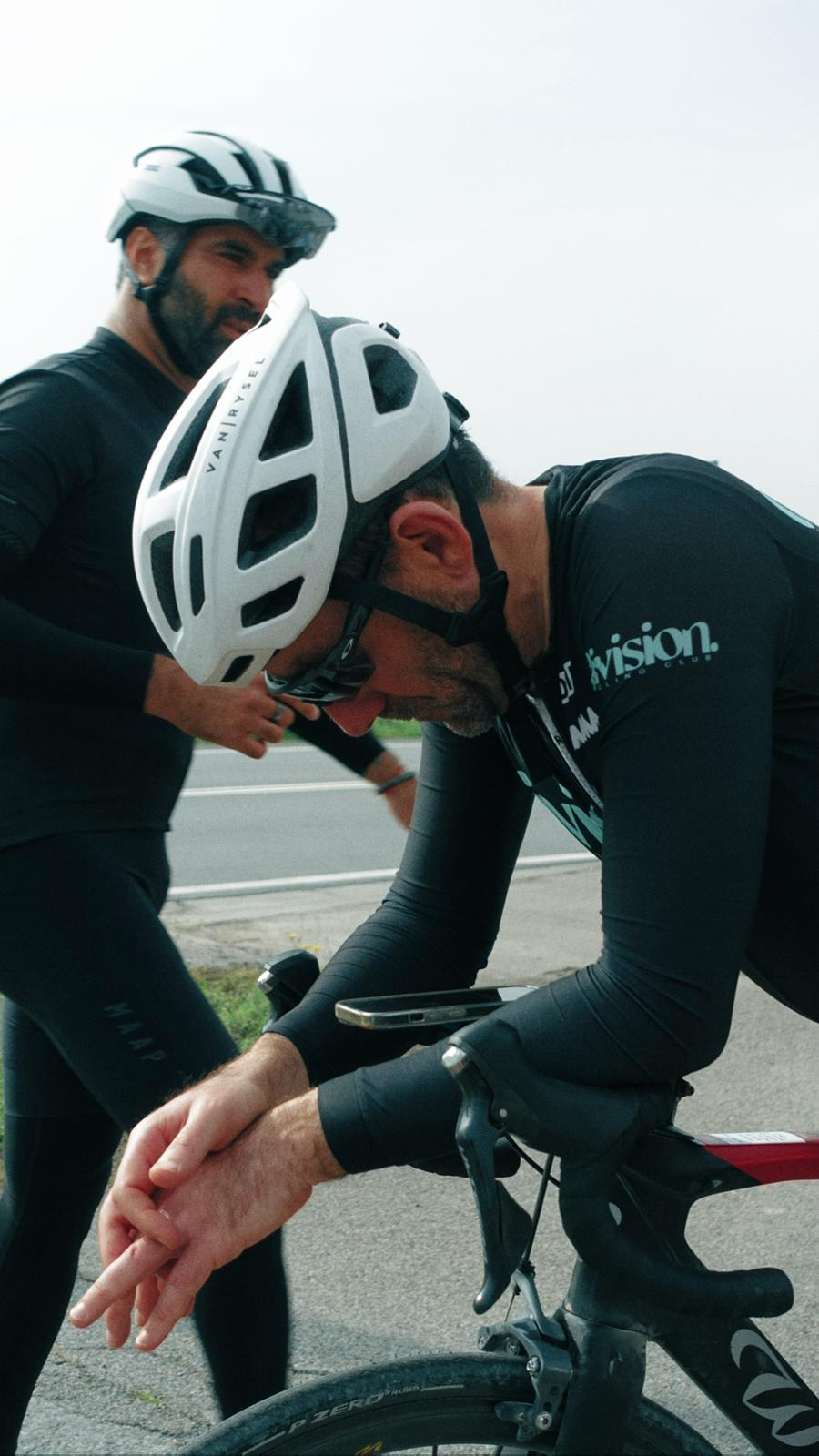 Dois ciclistas usando capacetes, um deles está abaixado no guidão da bicicleta enquanto o outro parece oferecê-lo suporte ou ajuda, ao lado de uma estrada.