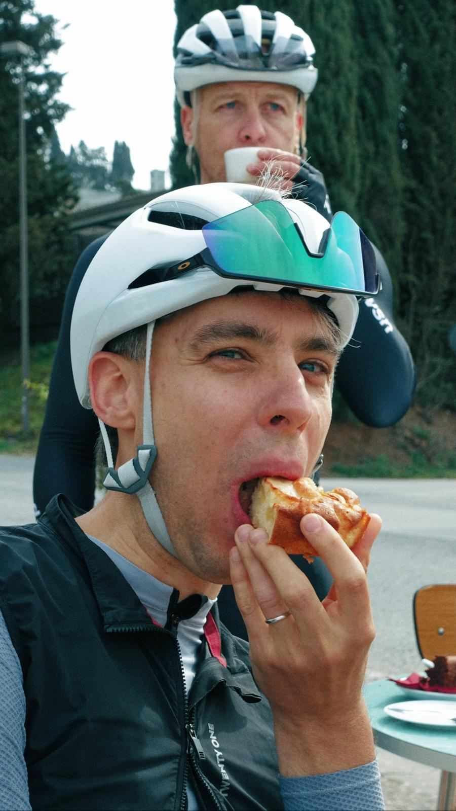Homens de bicicletas, um deles com capacete, tomando café e comendo pizza ao ar livre, com árvores ao fundo.