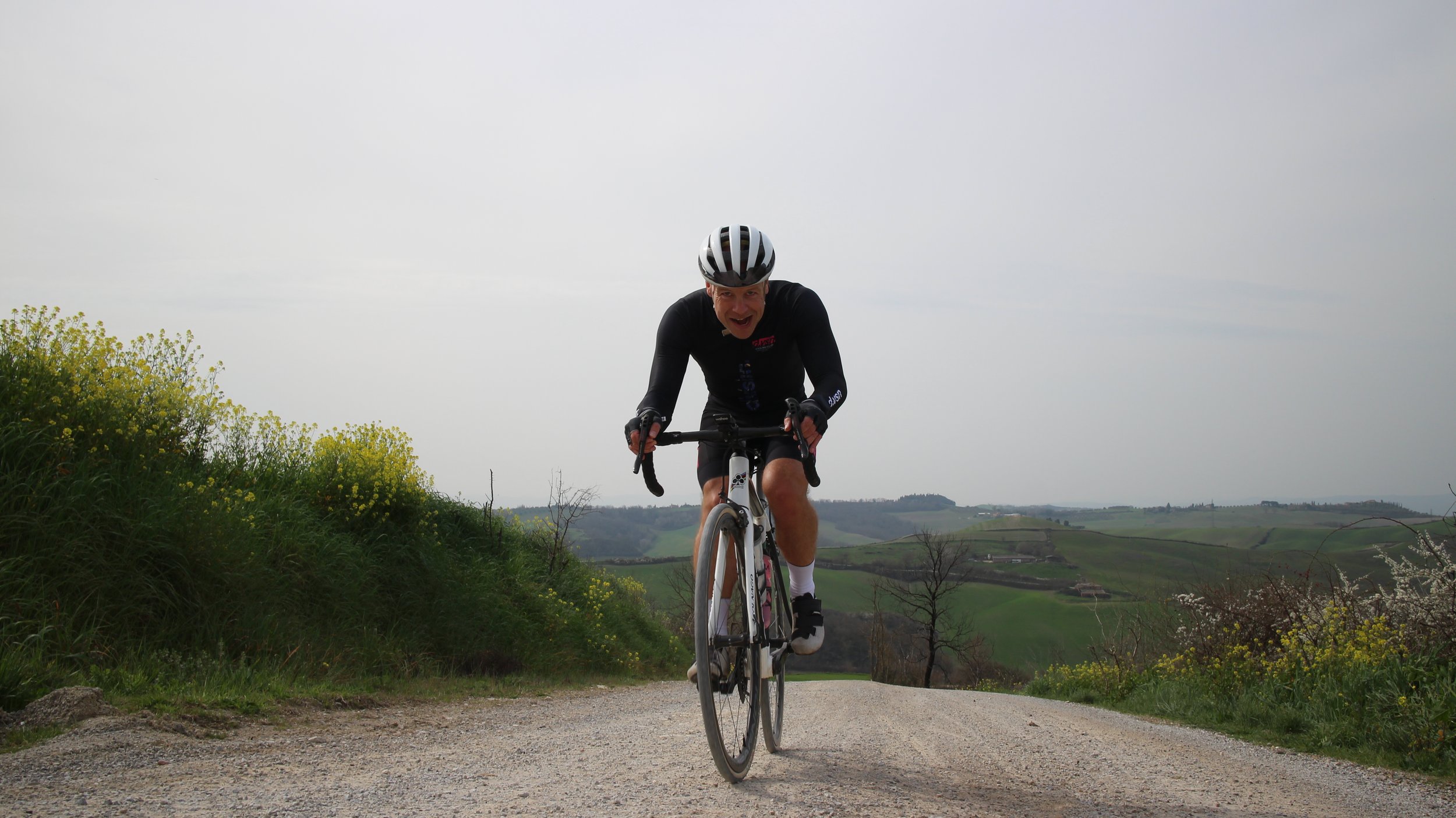 Homem sorridente pedalando de bicicleta em trilha natural com paisagem de colinas verdes ao fundo.