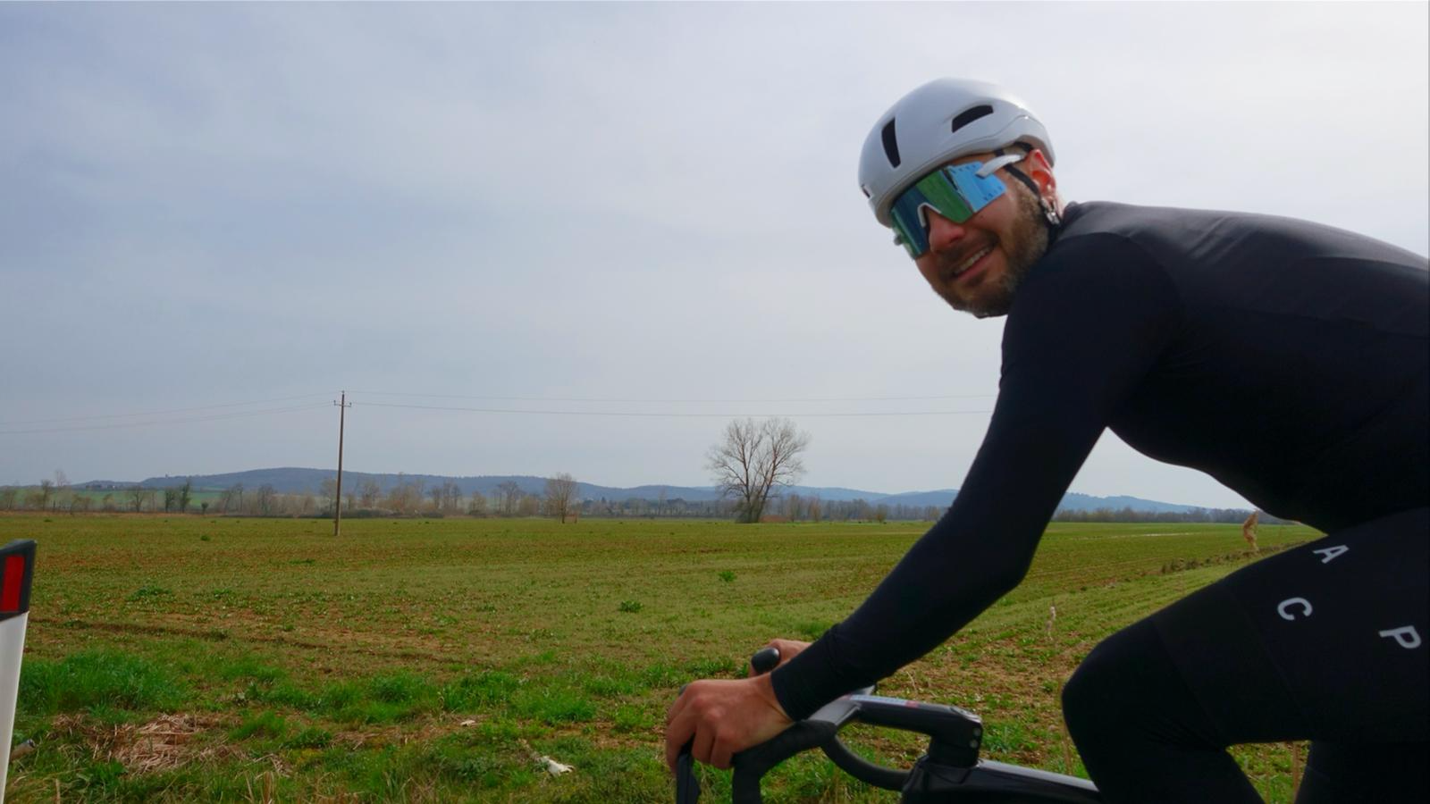 Homem alegre usando capacete e óculos de óculos de sol, pedalando de bicicleta em campo aberto com árvores e colinas ao fundo