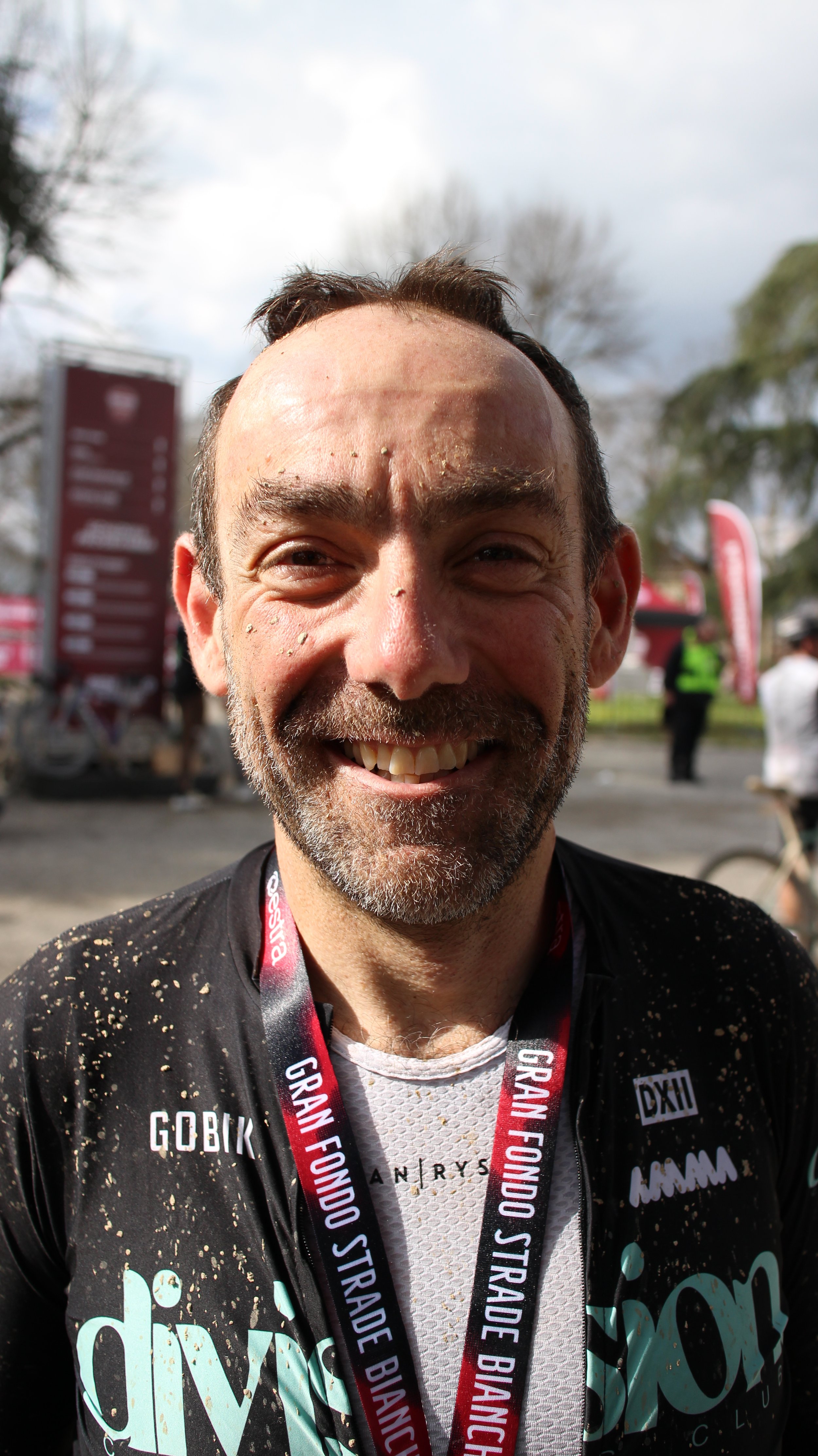 Homem sorridente com barba, vestindo camiseta e uma medalha de concorrer na corrida organizado pela Bianchini. O fundo mostra árvores, pessoas e banners em evento ao ar livre.