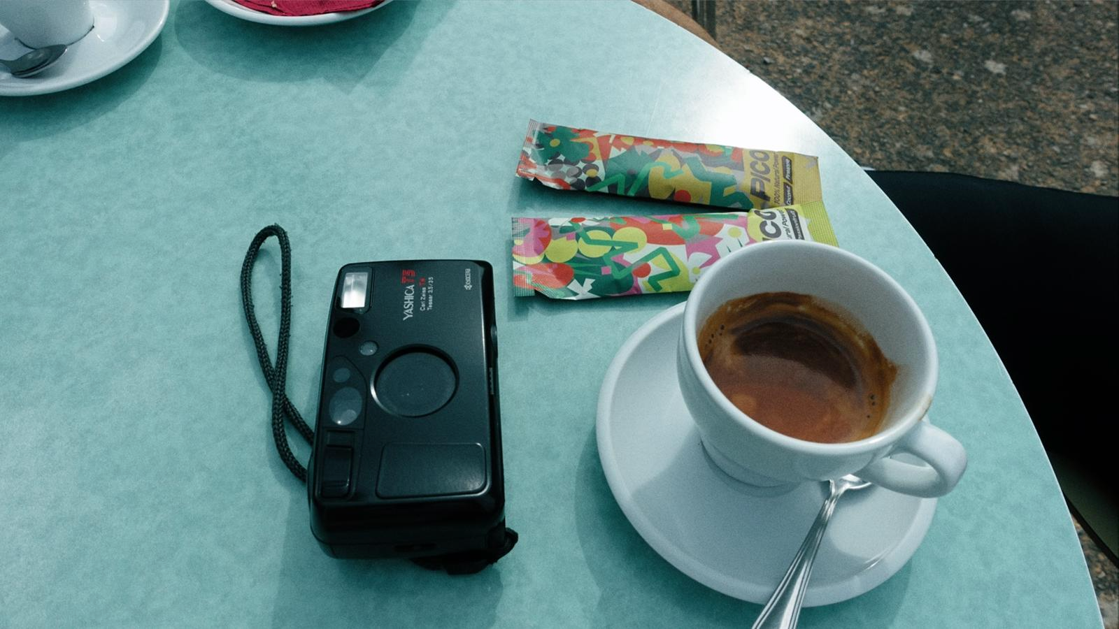 Tabela de mesa com uma xícara de café, uma câmera instantânea preta, dois chicletes coloridos e uma colher.