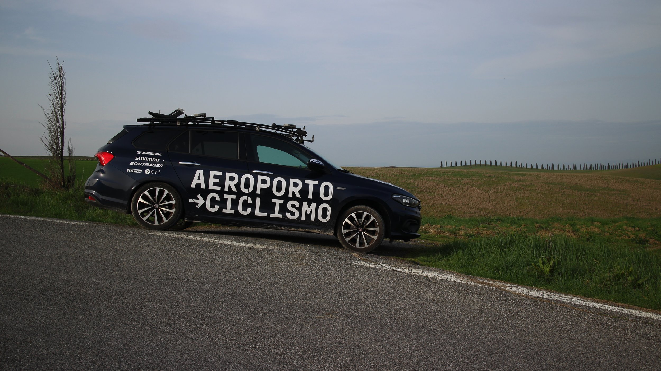 Carro preto estacionado ao lado de uma estrada, com a inscrição 'Aeroporto Ciclismo' em grandes letras brancas na lateral, em um campo aberto sob céu nublado.