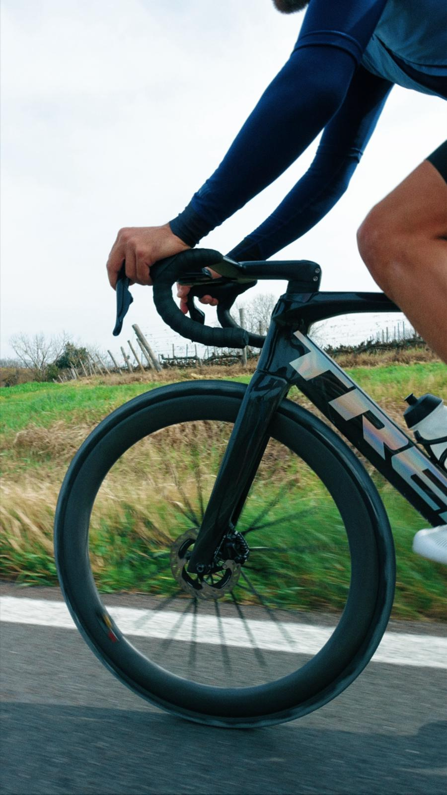 Pessoa pedalando uma bicicleta de estrada preta ao ar livre em uma estrada marcada com linha branca ao lado, com campo e árvores ao fundo em um dia nublado.