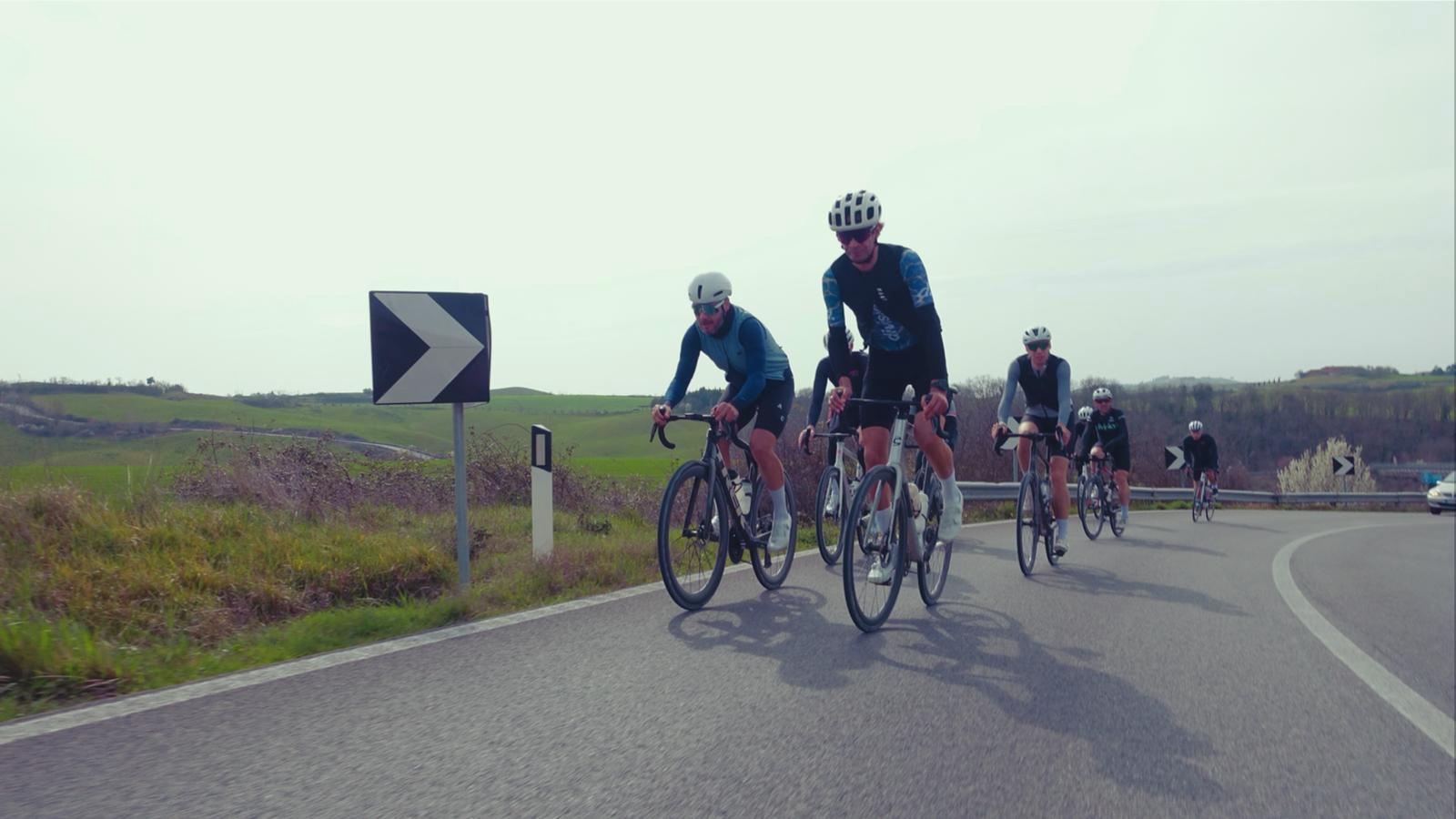 Grupo de ciclistas em uma curva de estrada rural, usando capacetes e roupas esportivas.