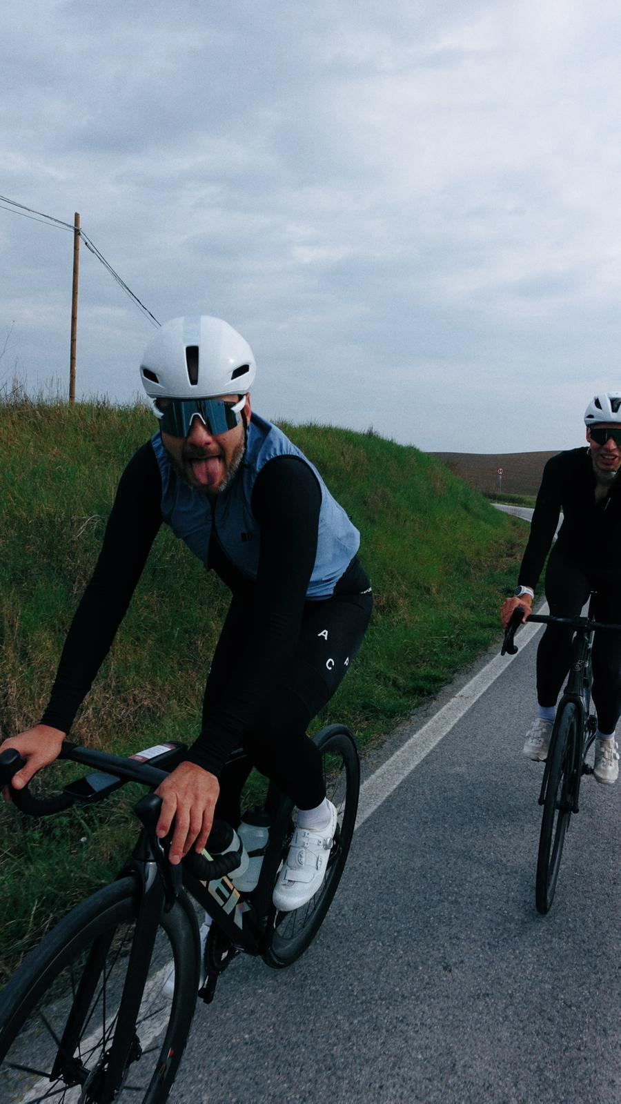 Dois ciclistas praticando bike de estrada em uma estrada rural, usando capacetes e óculos de proteção, com céu nublado ao fundo.