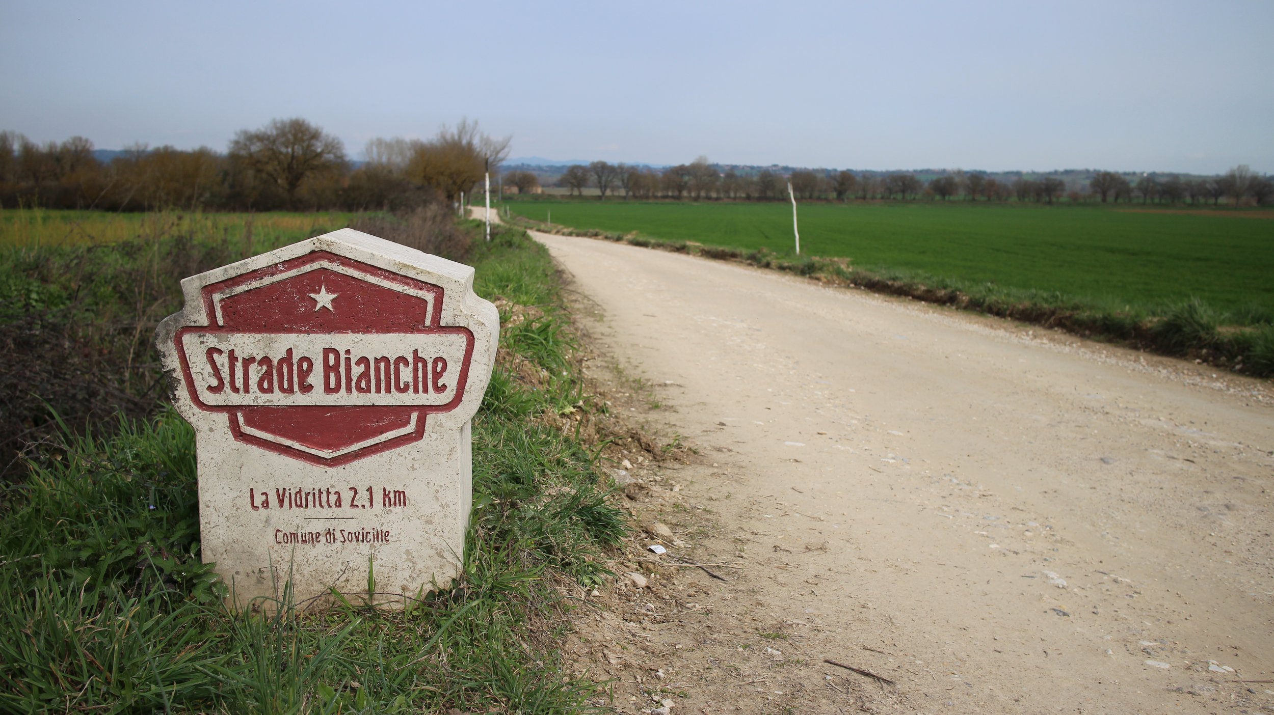 Placa de sinalização na beira da estrada com a inscrição 'Strade Bianche', indicando a distância de 2.1 km até La Vidritta, em uma área rural com estrada de terra, campos verdes e árvores ao fundo.