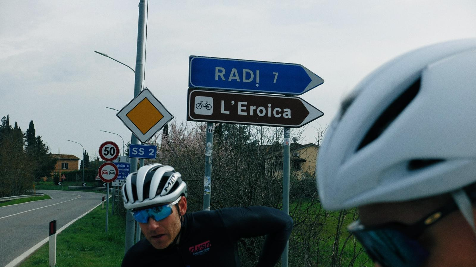 Ciclistas com capacetes na estrada, sinais de trânsito ao fundo, incluindo placas de limites de velocidade e direções em italiano, cercado por árvores e casas rurais.