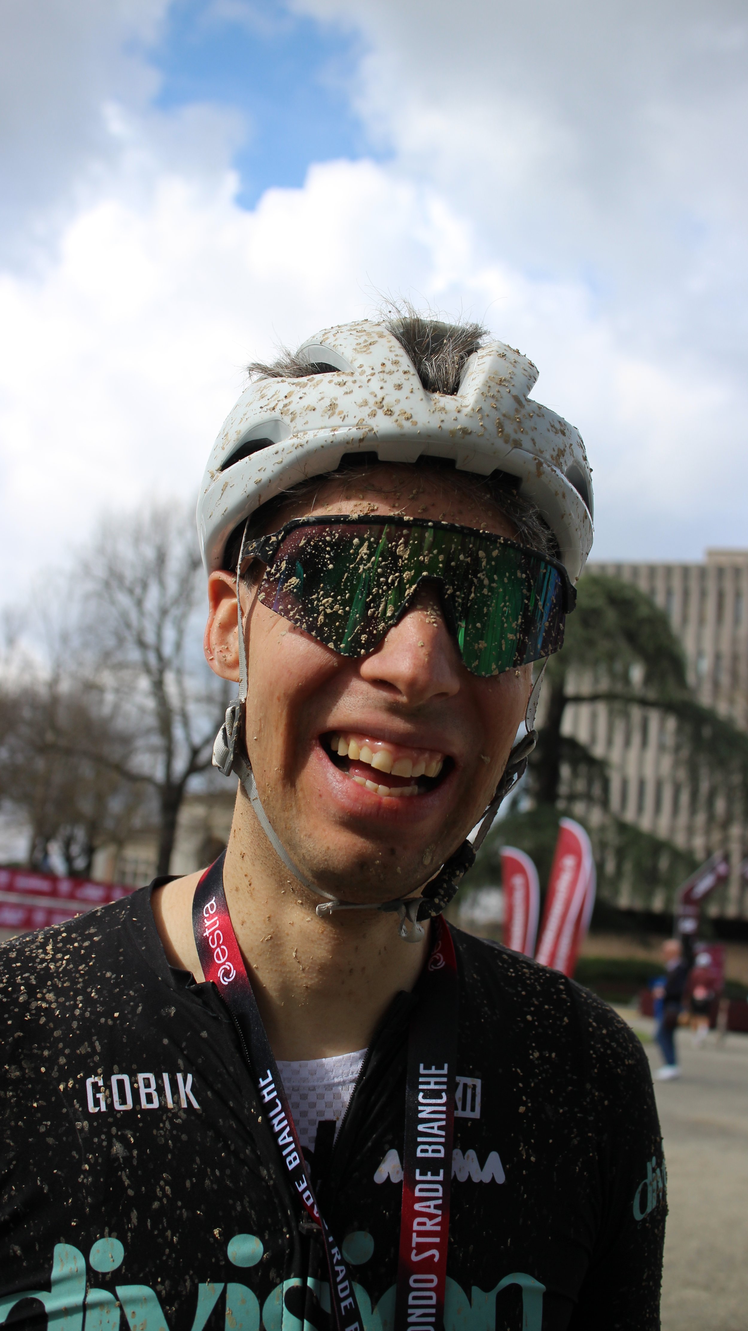 Pessoa sorrindo vestida com roupa de ciclismo, usando capacete e óculos de proteção, coberta de lama, ao ar livre, após uma atividade de ciclismo.