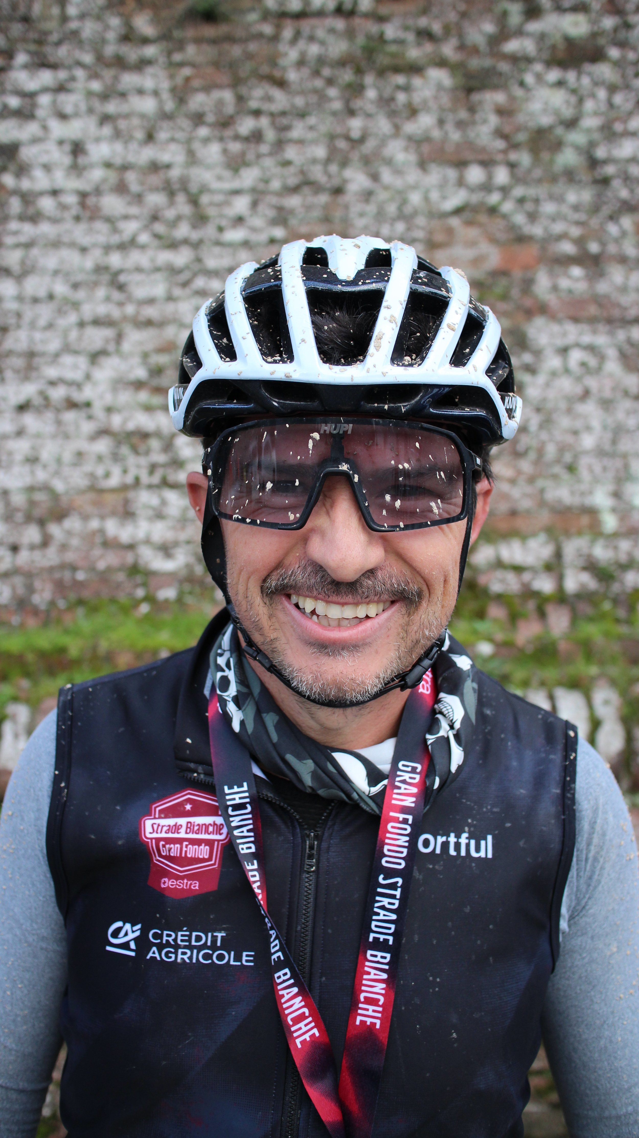 Homem sorrindo vestindo capacete de ciclismo, óculos de proteção, jaqueta esportiva e medalhas, posando na frente de uma parede de tijolos.