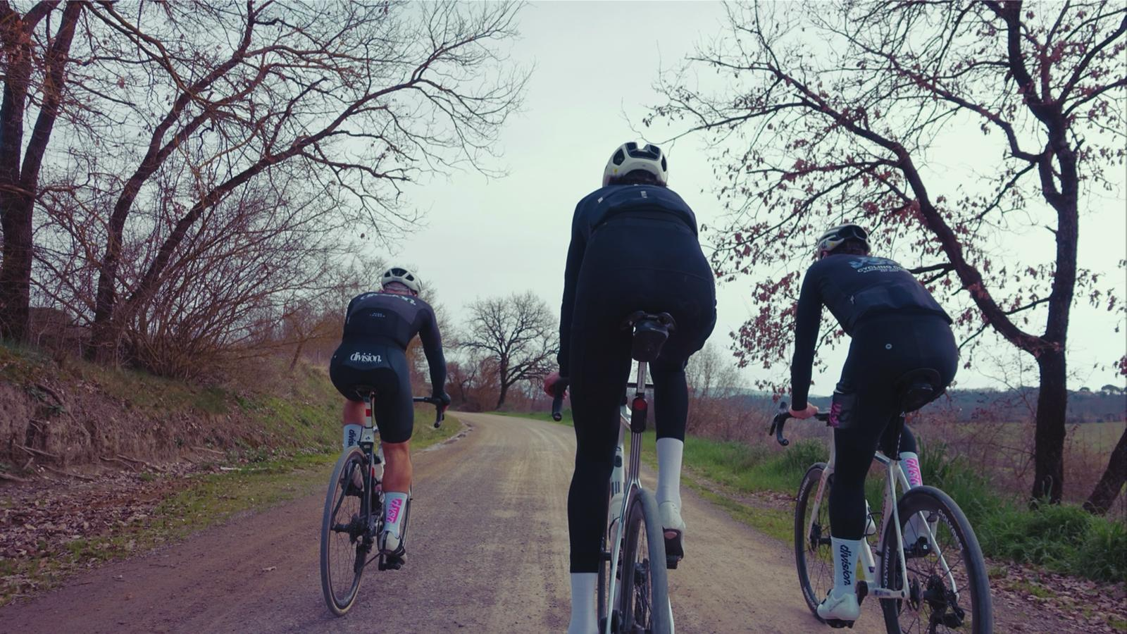 Três ciclistas pedalando em uma estrada de terra, rodeados por árvores nuas, em clima de fim de tarde ou início de manhã.