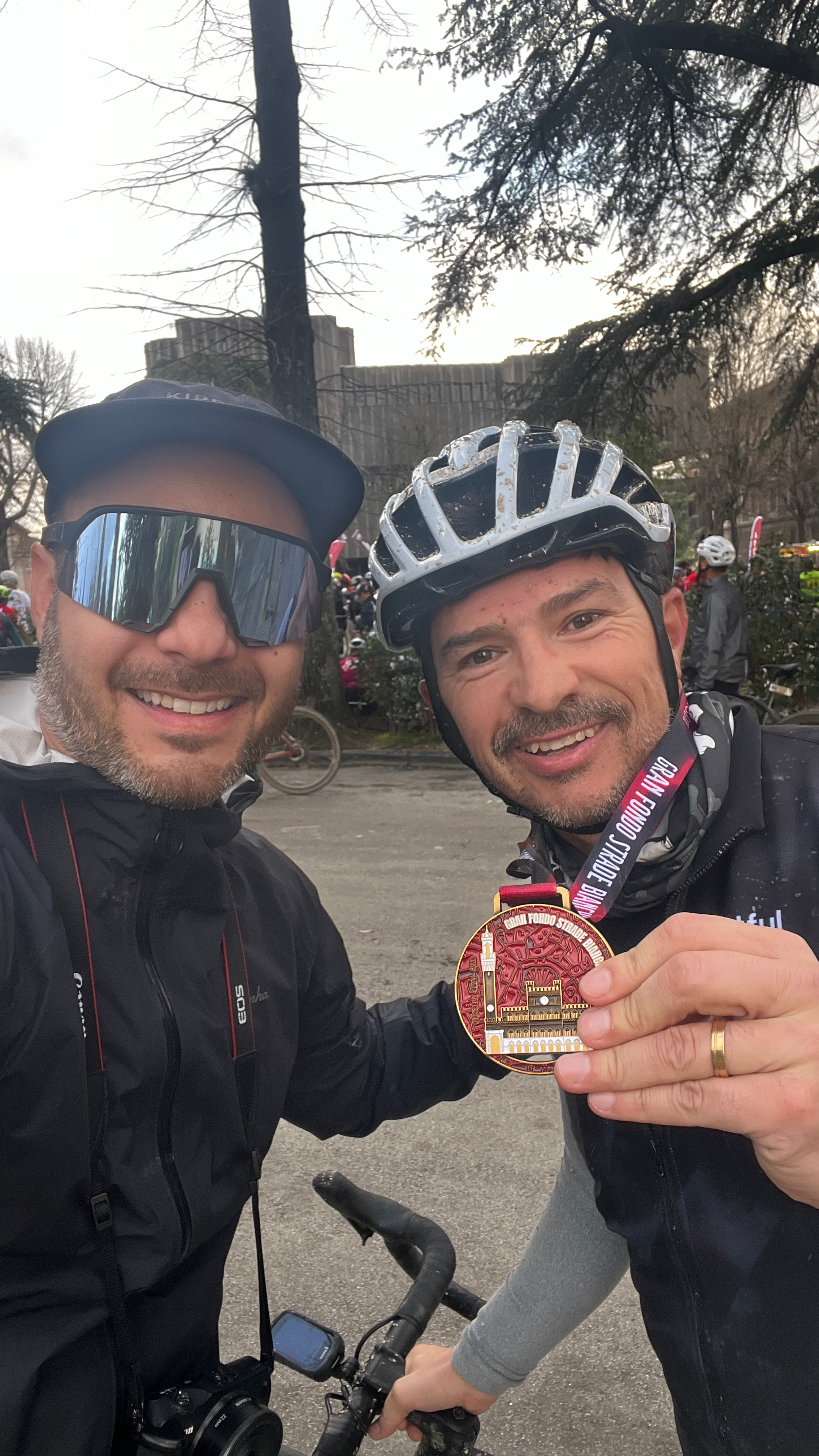 Dois homens sorrindo, um segurando uma medalha e o outro com uma câmera ao redor do pescoço, ambos usando capacetes de bicicleta, em um evento ao ar livre.