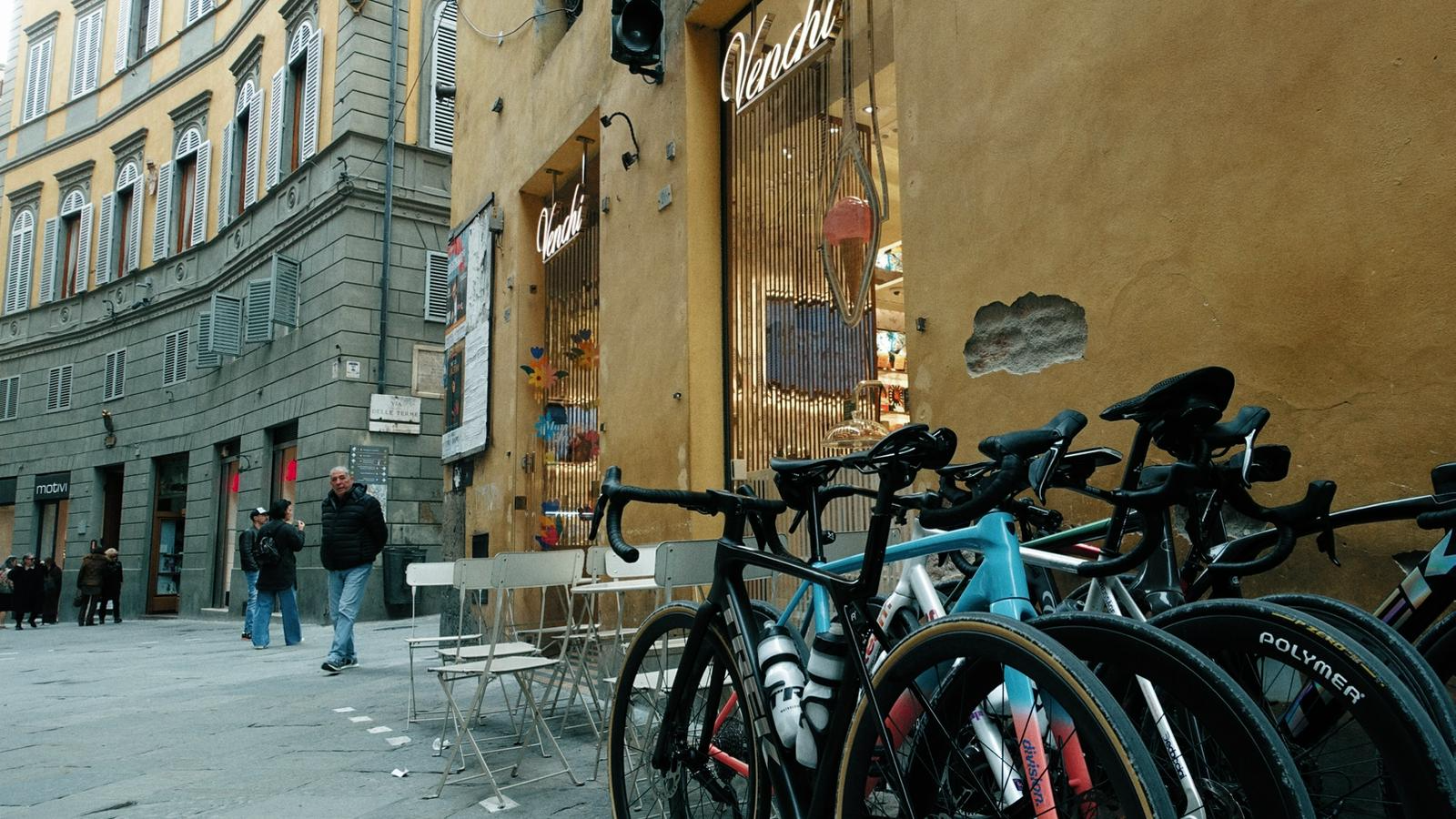 Rua com prédios de arquitetura clássica, várias bicicletas alinhadas na calçada, pessoas caminhando, uma loja de doces chamada 'Vadel' com uma fachada amarela.