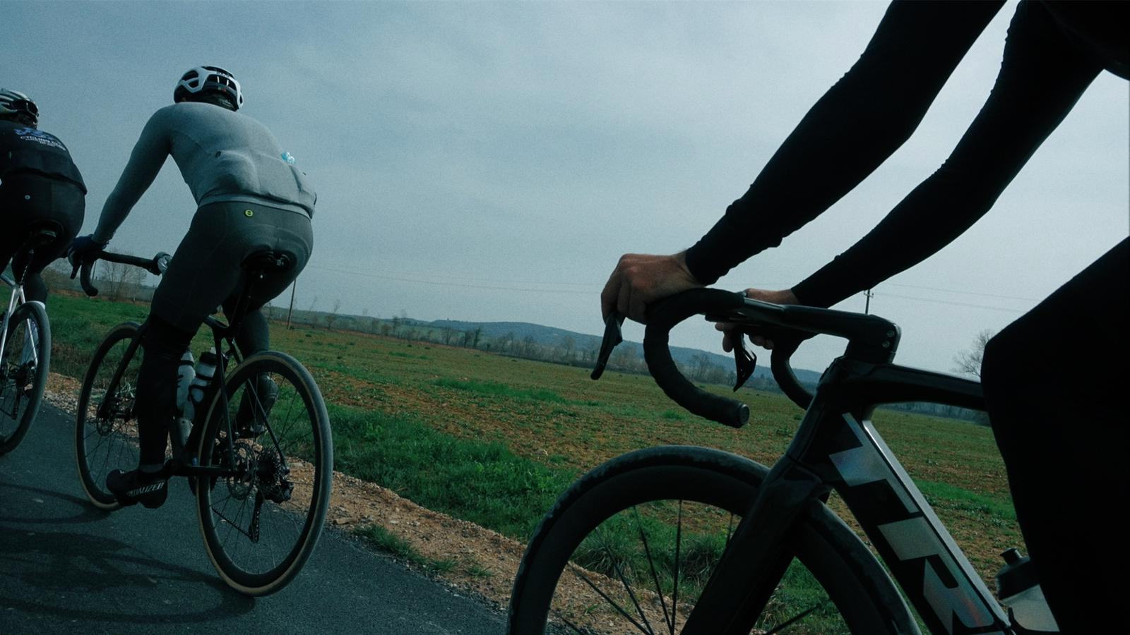Três ciclistas pedalando em uma estrada no campo sob céu nublado, com vegetação e árvores ao fundo.