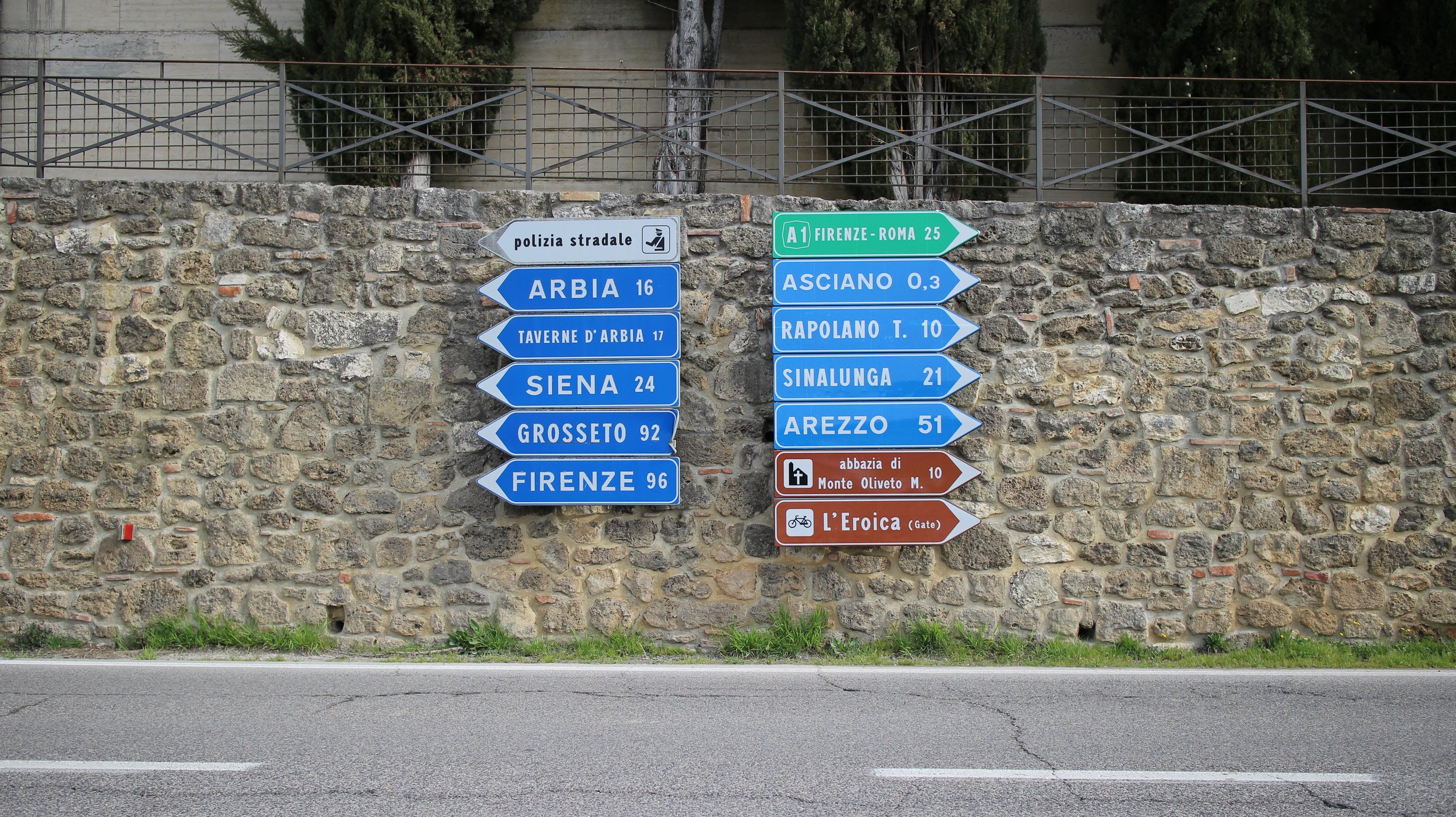 Placas de trânsito em uma parede de pedra, indicando direções para várias cidades italianas e pontos turísticos, com uma cerca de metal ao fundo.
