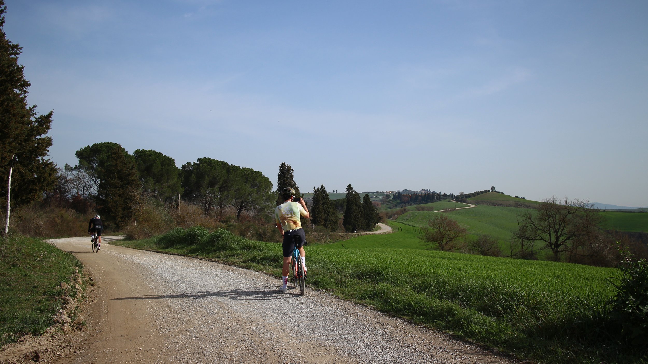 Ciclistas pedalando por uma estrada de terra em uma paisagem rural com colinas verdes, árvores e céu claro.