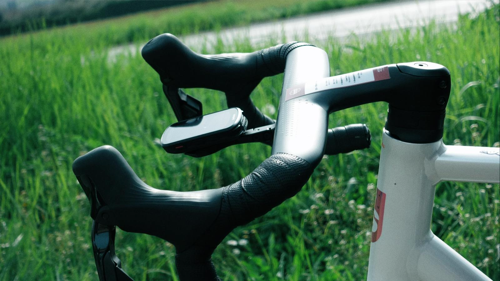 Detalhes do guidão de uma bicicleta de estrada com quadro branco, configurada para corrida ou ciclismo de alta performance, com fundo de campo verde.