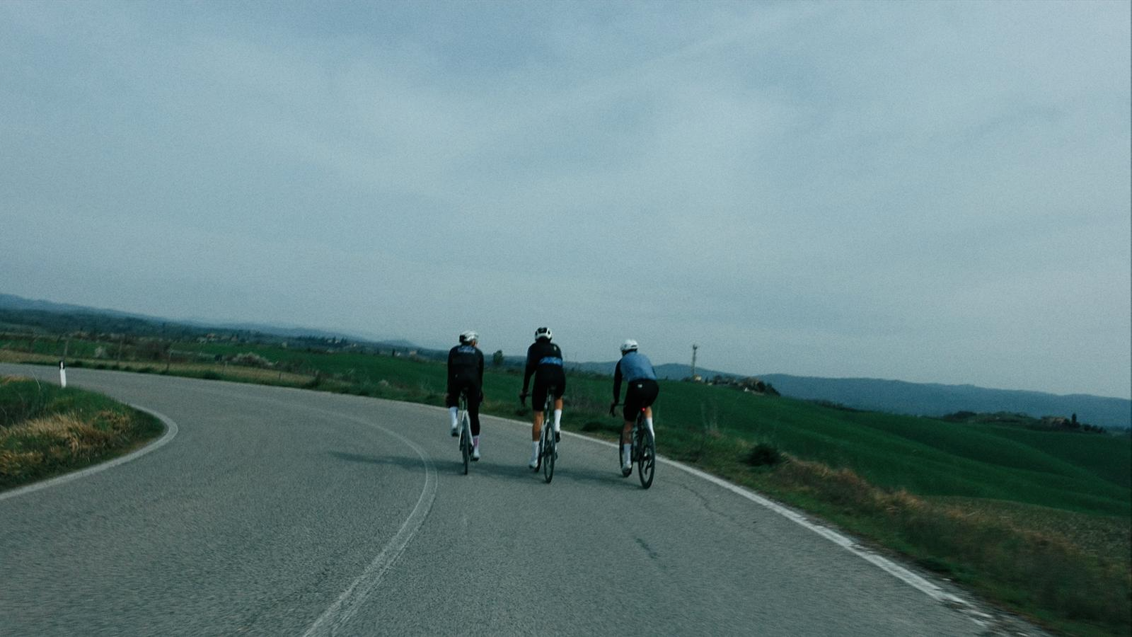 Três ciclistas pedalando em uma estrada rural cercada por campos verdes, sob céu nublado.