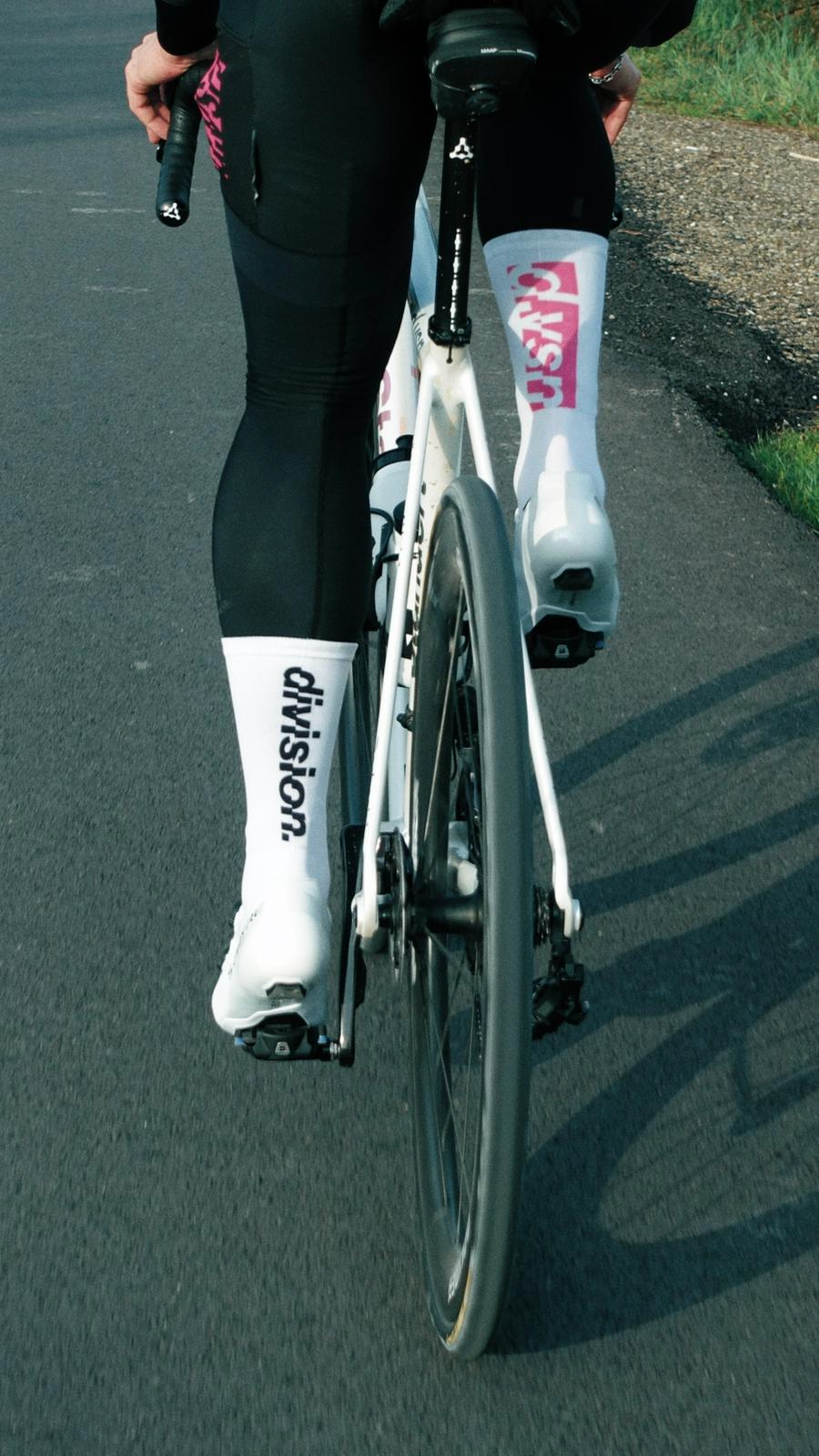 Pessoa andando de bicicleta na estrada, vestindo roupas esportivas brancas e pretas, com meias brancas e o nome 'JUSTDOIT' impresso nelas.
