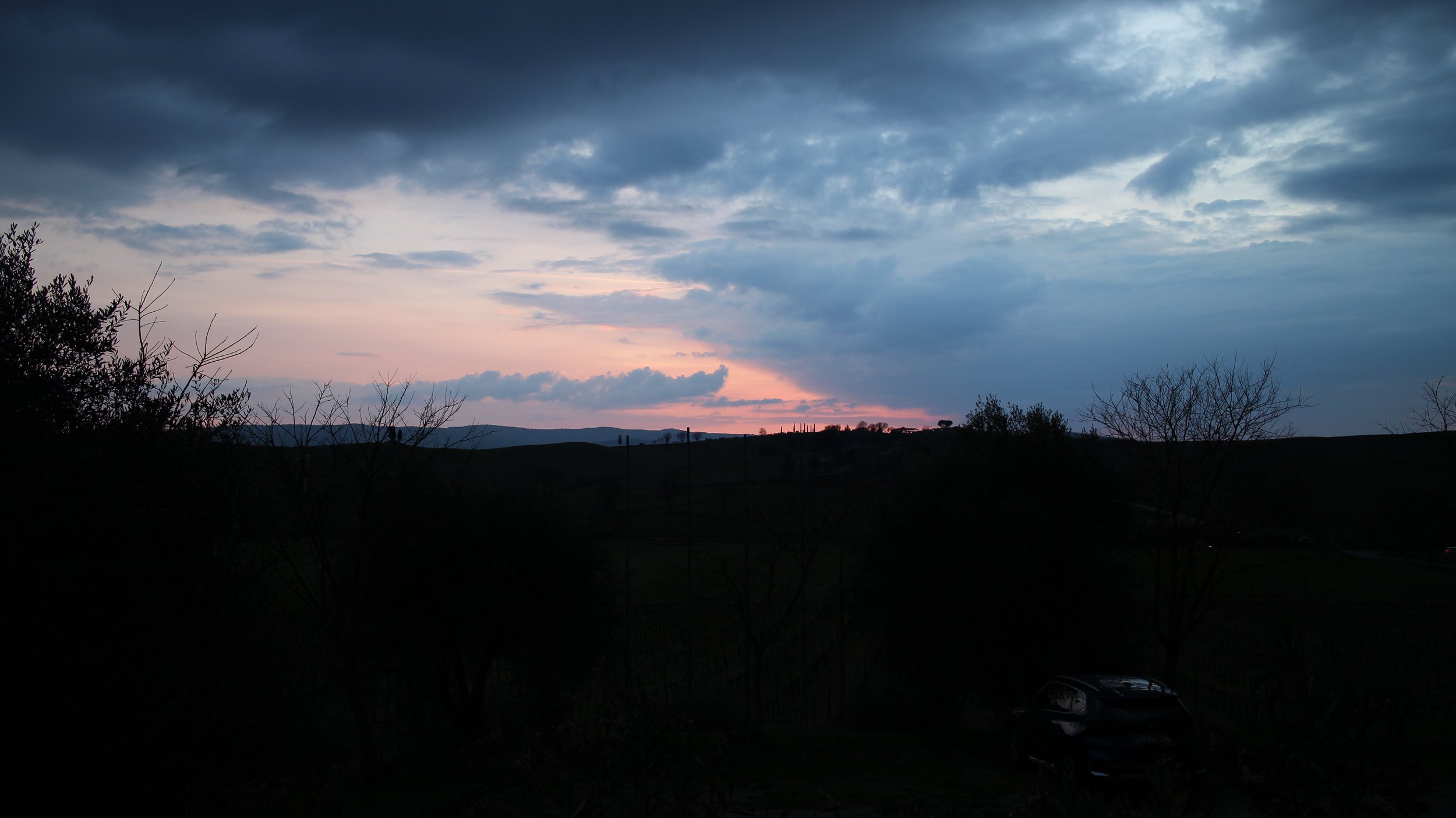 Céu ao entardecer com nuvens escuras, árvores ao fundo e uma colina ao horizonte, com um carro estacionado na parte inferior direita.