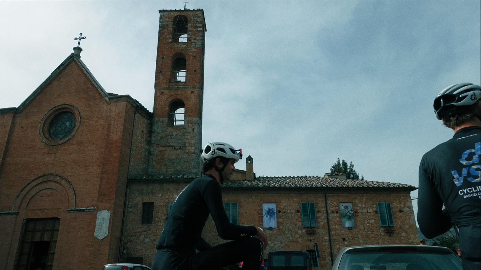 Homens de bicicleta descansando em frente a uma igreja de estilo antigo com torre de sino de tijolos e janelas de coruja, em um dia nublado.