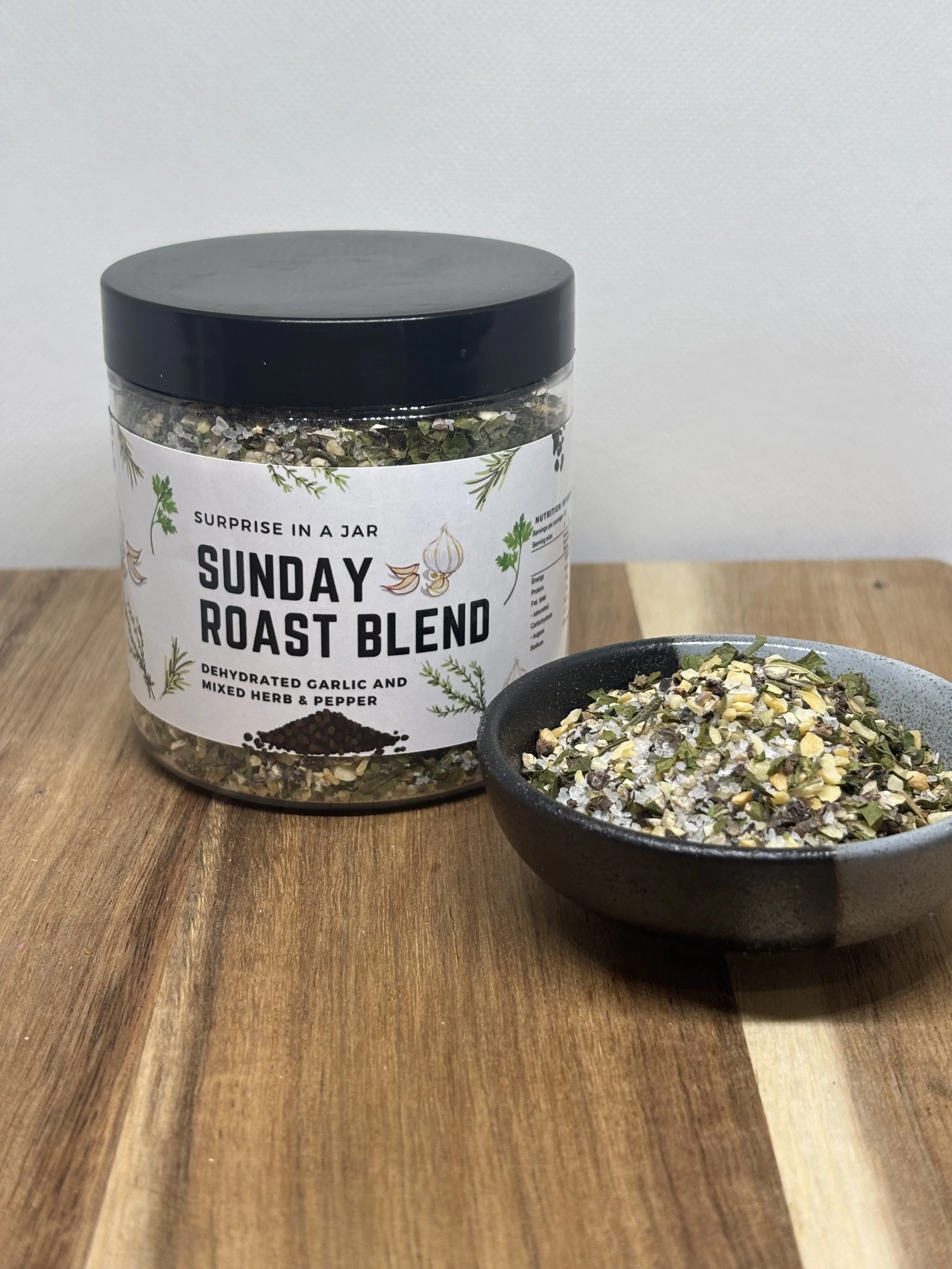 A jar labeled "Sunday Roast Blend" and a black bowl containing a mixture of dried herbs and pepper on a wooden surface.