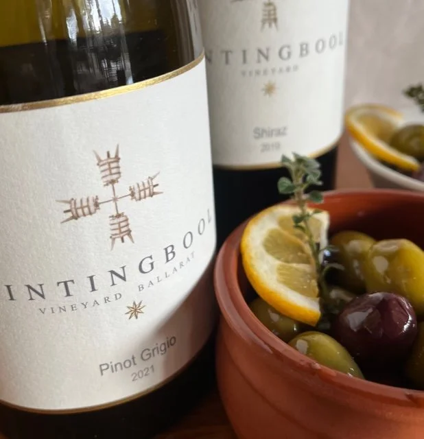 Two bottles of Tingboo Vineyard Ballarat Pinot Grigio 2021 wine and a terracotta bowl filled with green and black olives garnished with lemon slices and a sprig of herbs.