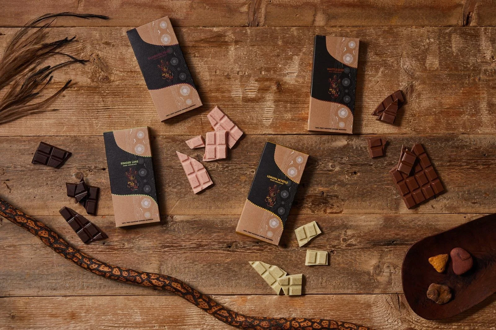 Assorted chocolates in different flavors and types placed on a rustic wooden surface, with some chocolates in their wrappers and others unwrapped, along with a wooden dish holding additional chocolates, a corn husk, and dried plants.