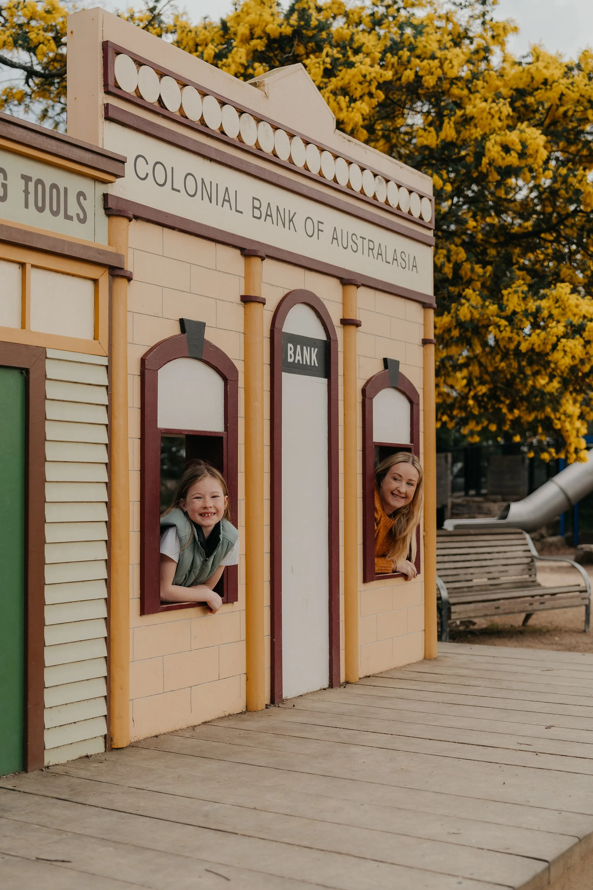 A girl and a woman, looking out of window of small colourful playhouse with a train station theme, titled "Colonial Bank of Australasia," in park with benches and trees with yellow flowers.