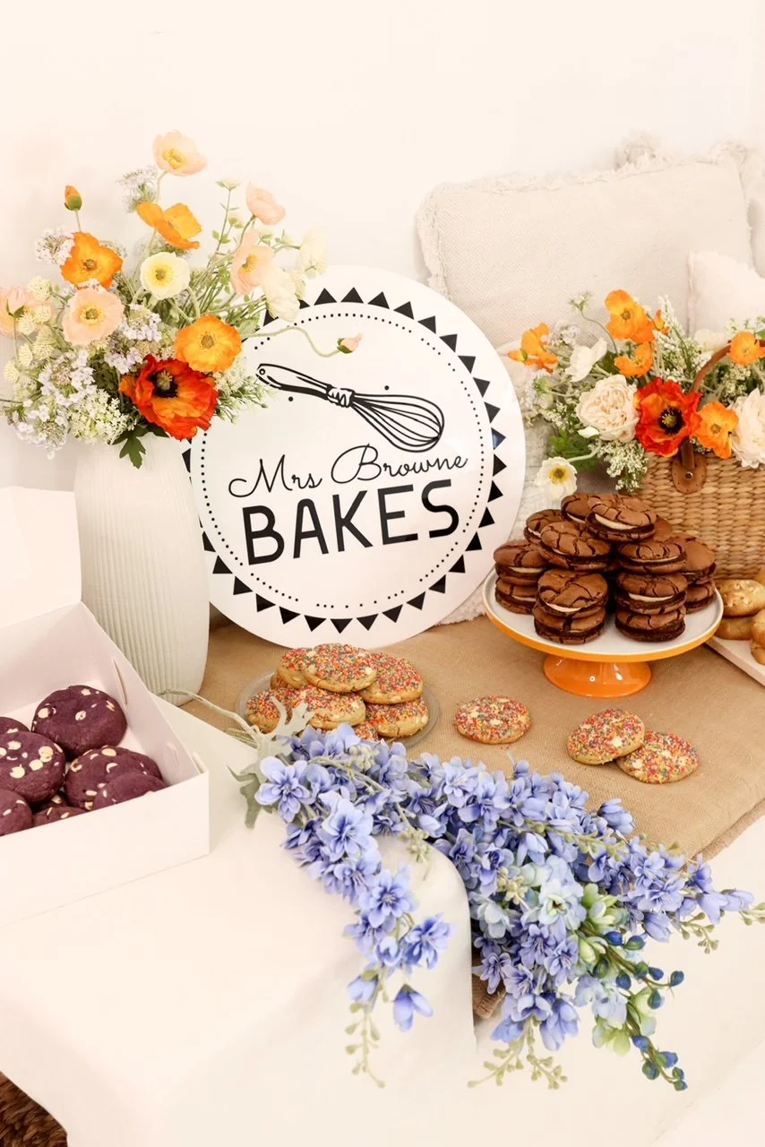 Table decorated with colorful flowers, cookies, and chocolates, with a sign reading 'Mrs. Browne Bakes' and a whisk illustration.