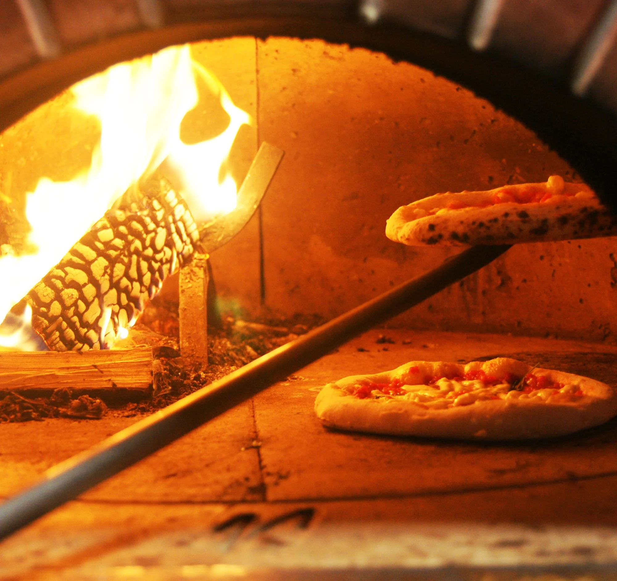 A pizza in an oven with flames visible and a pizza peel nearby.
