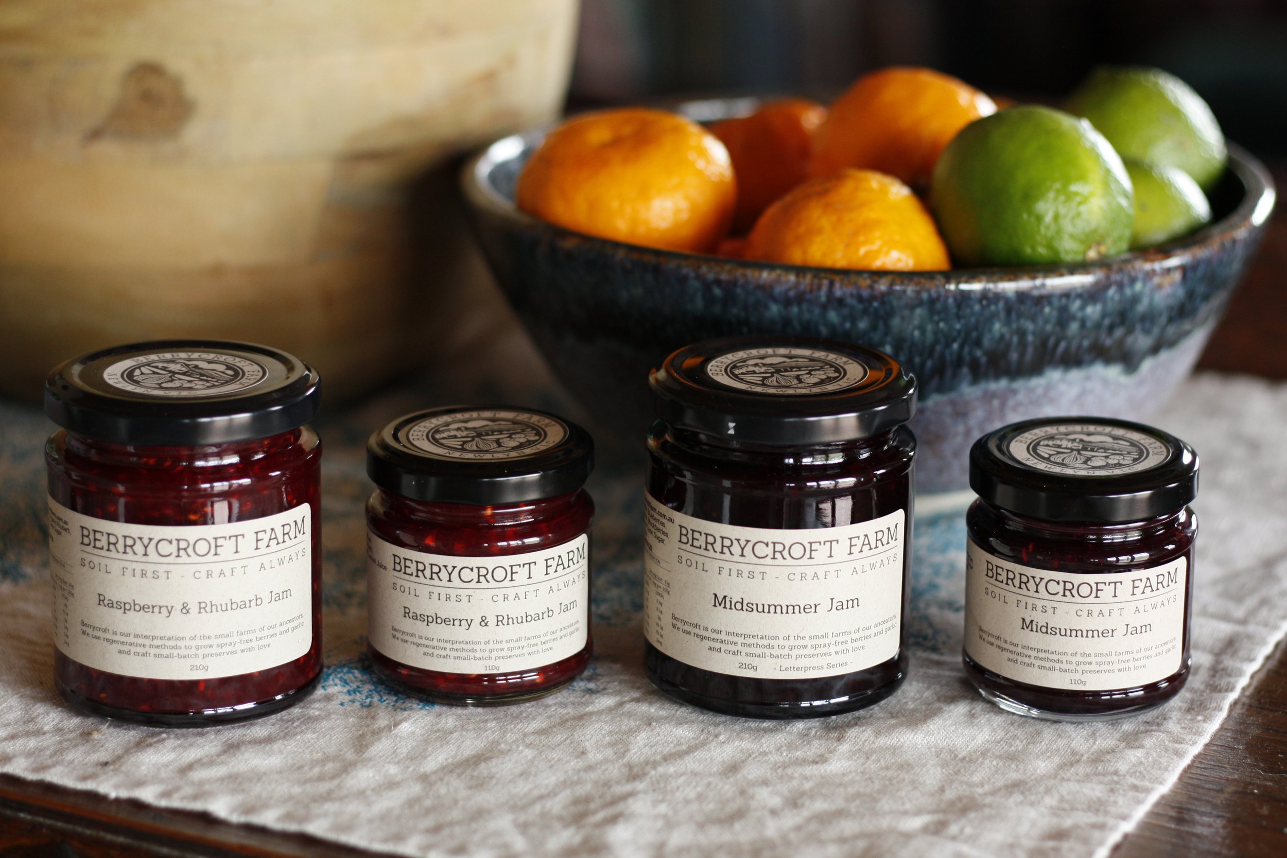 Four jars of berry jam with labels from Berry Croft Farm, placed on a cloth surface, with a bowl of oranges and limes in the background.