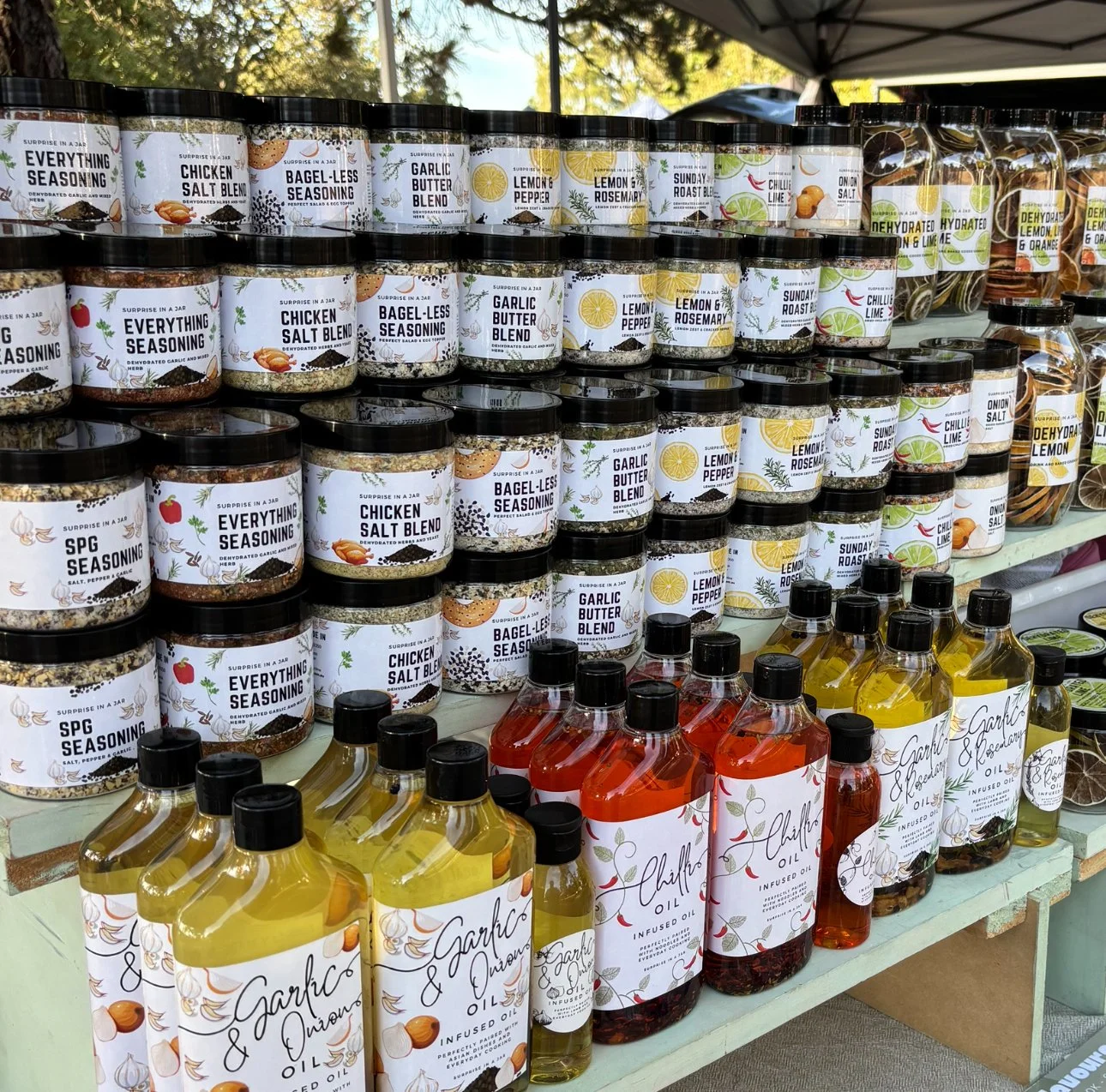 Display of various jars of seasoning and infused oils on a multi-tiered shelf at an outdoor market, with labels indicating different flavors and ingredients.