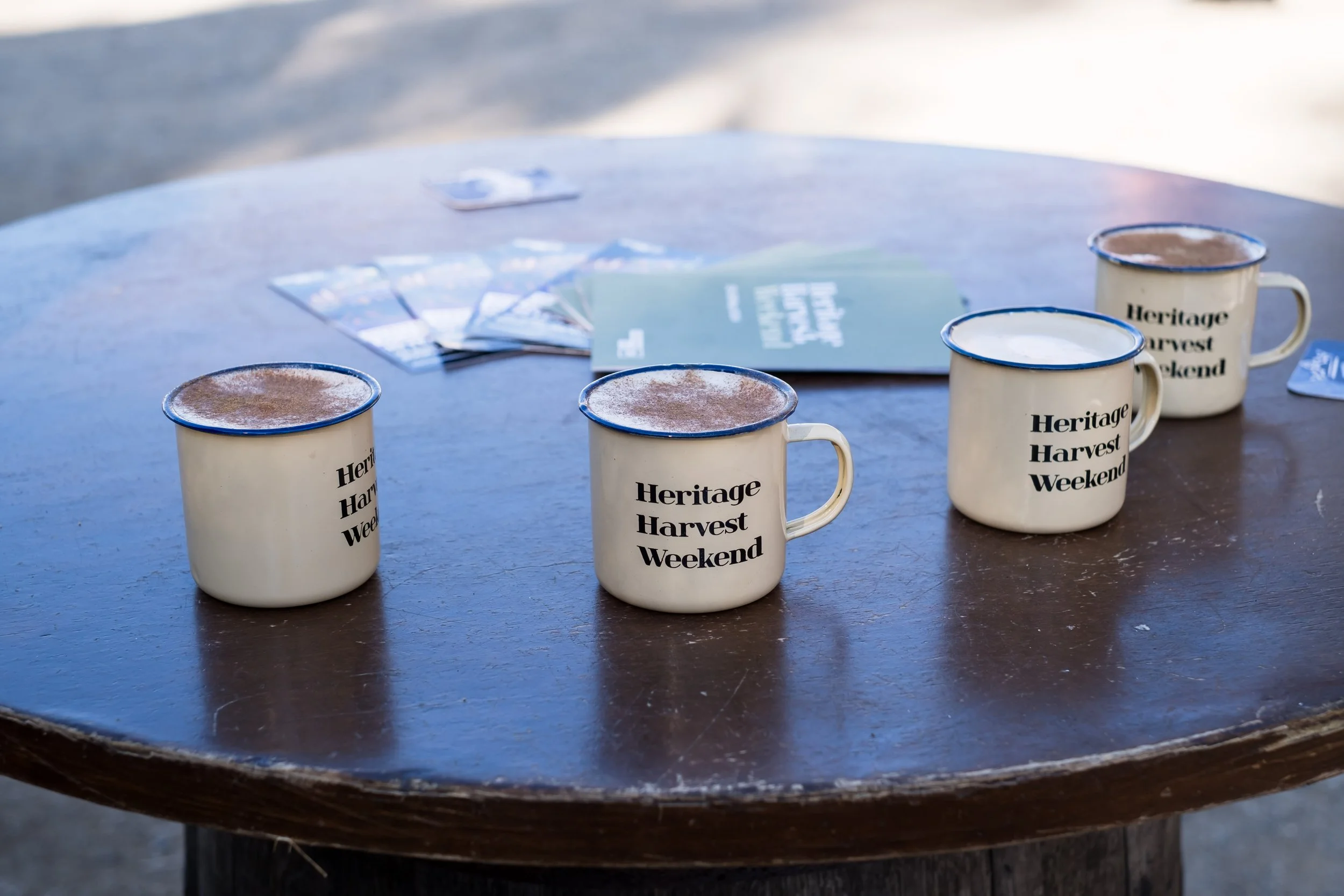 Four enamel mugs with the text 'Heritage Harvest Weekend' printed on them sit on a dark wooden table, each filled with a hot beverage, possibly coffee or hot chocolate, outside. In the background, there are brochures and brochures on the table.