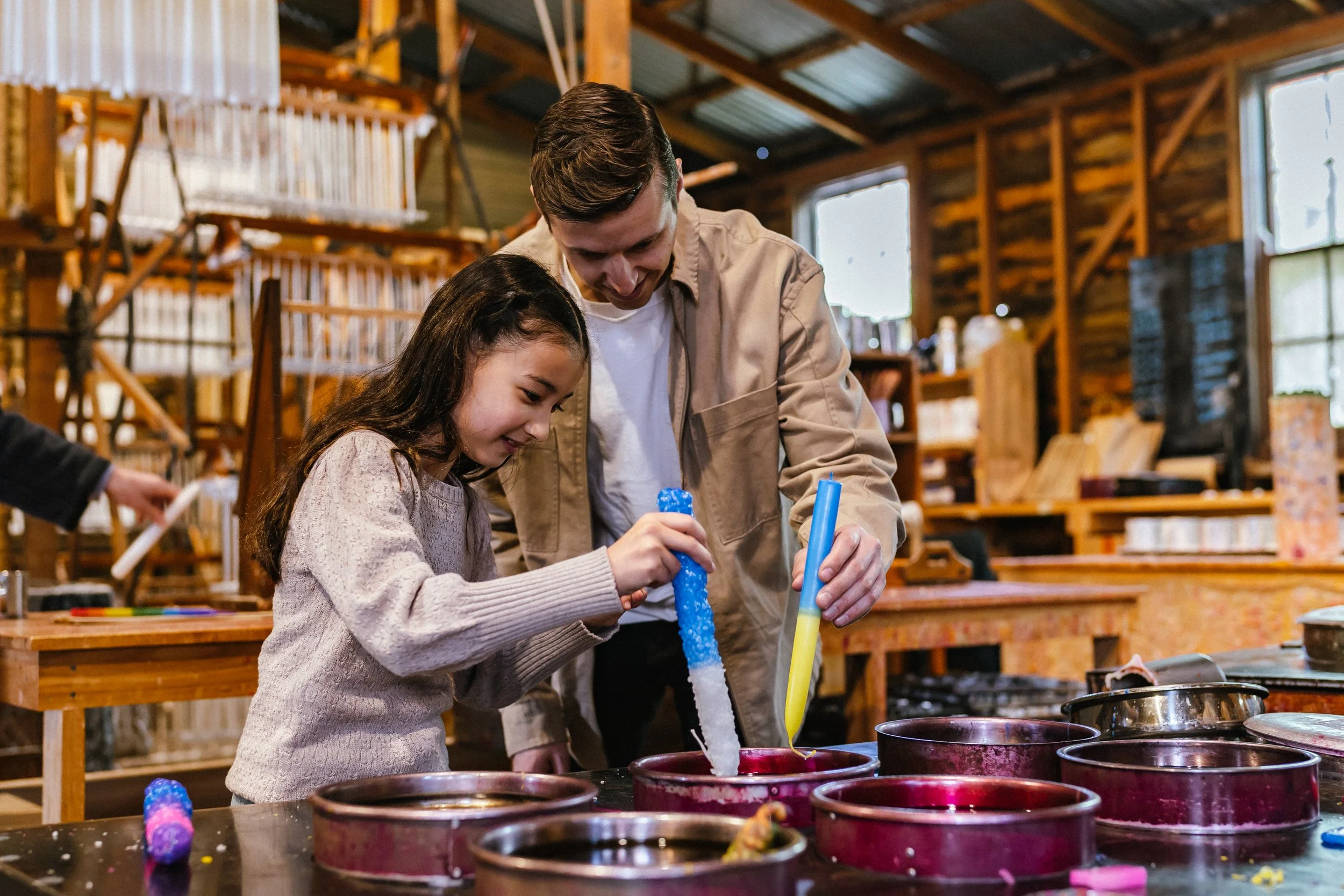 A man and a young girl are in a woodworking or craft workshop, working together on a project with colorful materials, surrounded by shelves of wood and craft supplies.