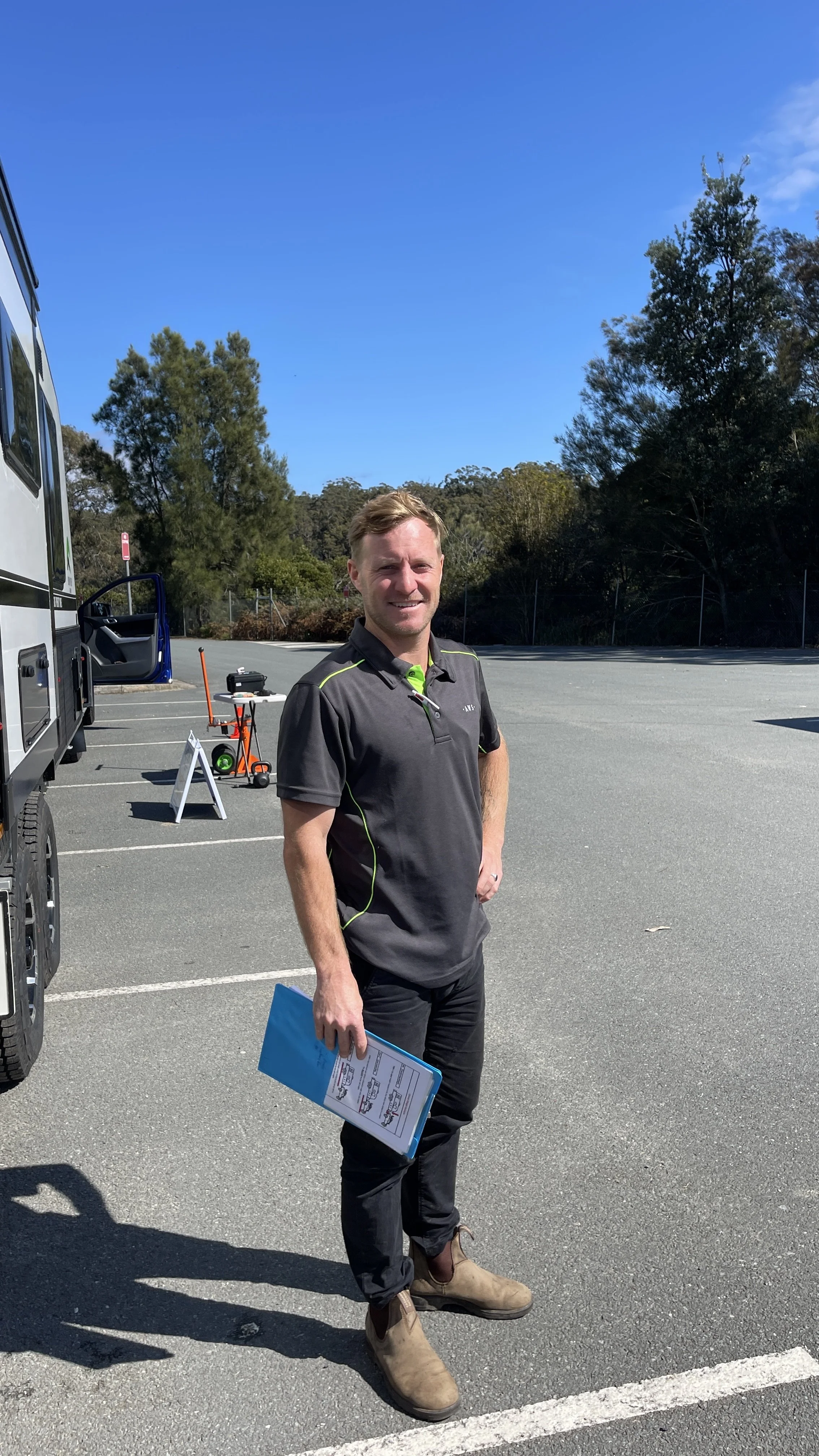 Man holding a clipboard in a parking lot next to a vehicle, under a clear blue sky with trees in the background.