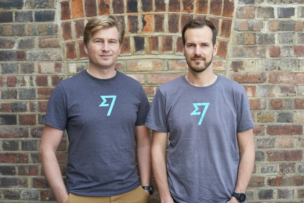Two men standing side by side against a brick wall, wearing matching gray T-shirts with a blue logo on the front.