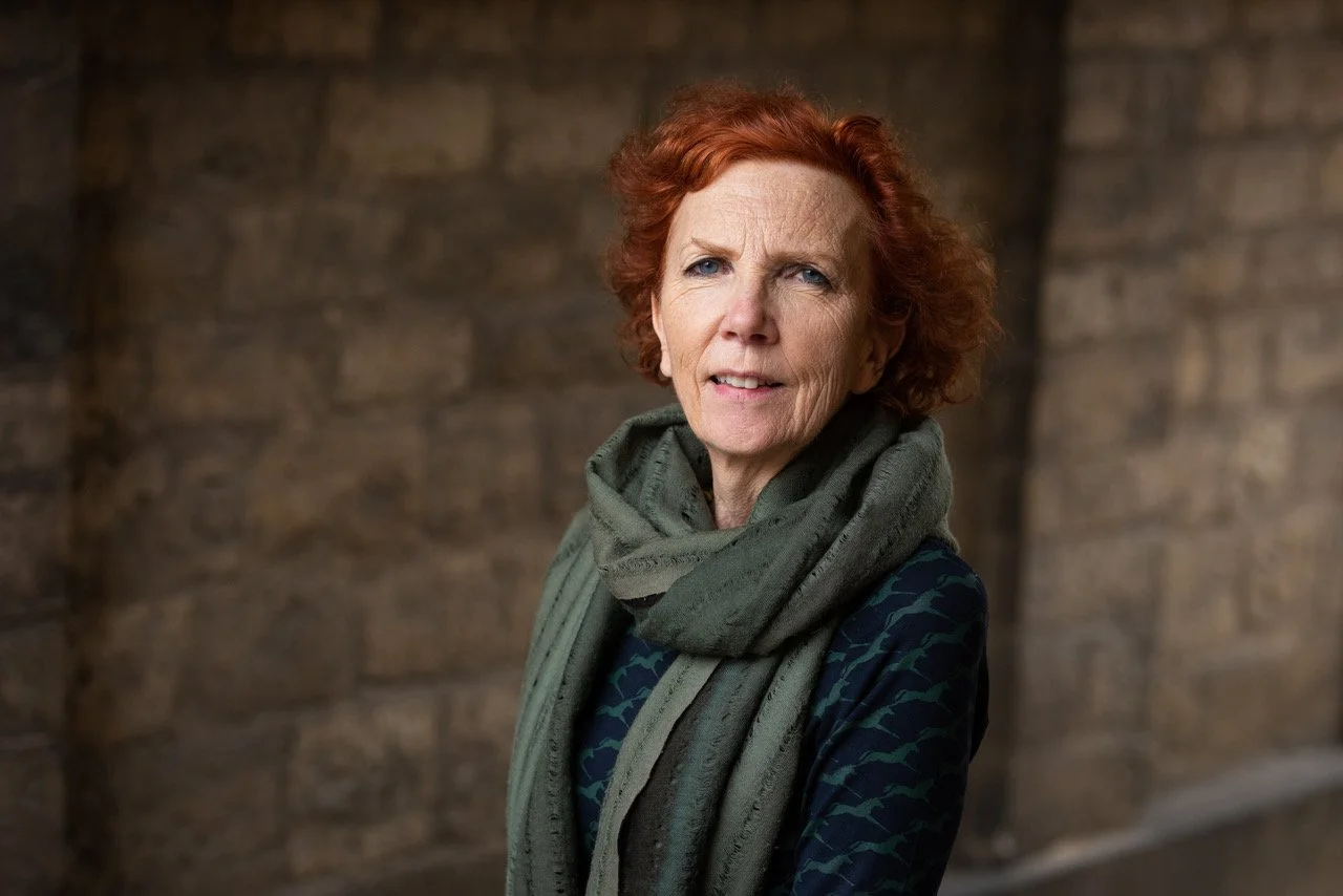 A woman with short red hair and blue eyes standing outdoors near a brick wall, wearing a green scarf and a dark patterned jacket.